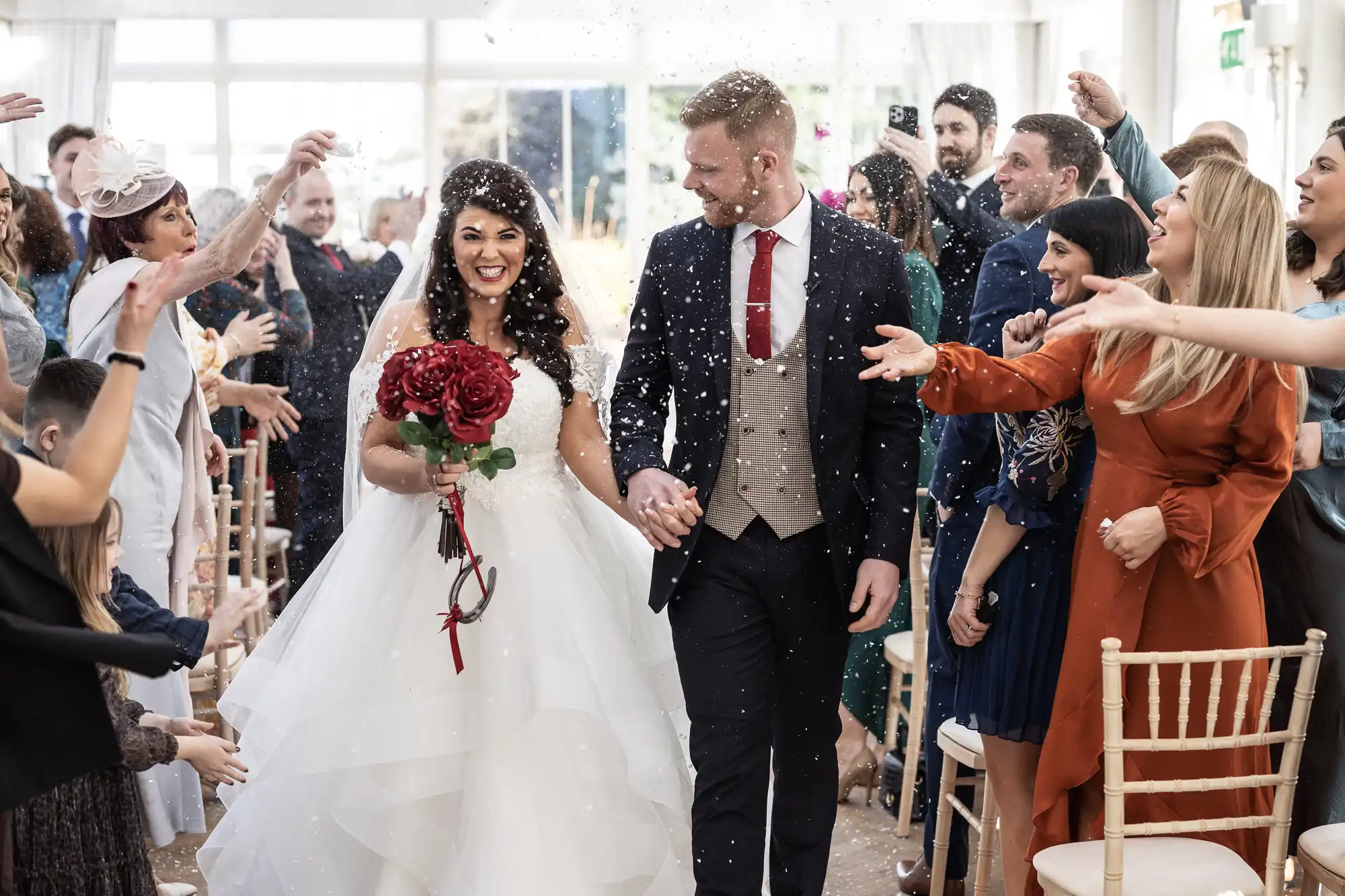 A bride and groom walk down an aisle while guests toss confetti. The bride holds a red bouquet, and both are smiling. Guests on either side are celebrating and taking photos.