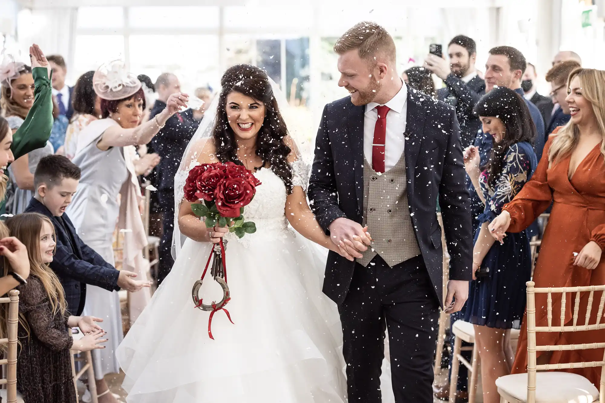 A bride and groom walk down the aisle holding hands, surrounded by guests throwing confetti. The bride carries a bouquet of red flowers.