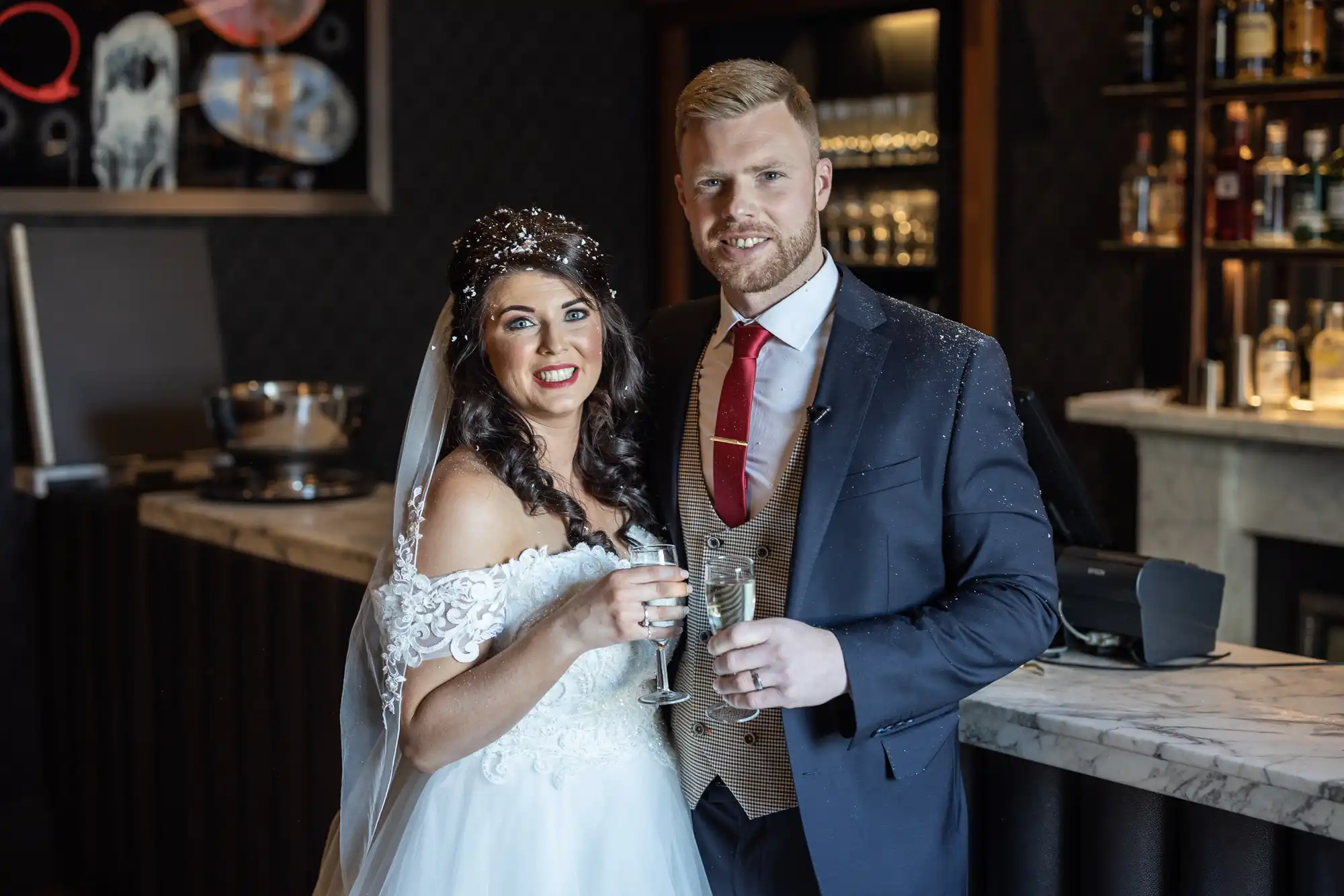 Bride in white dress and groom in a suit pose together holding glasses of champagne in a bar setting.