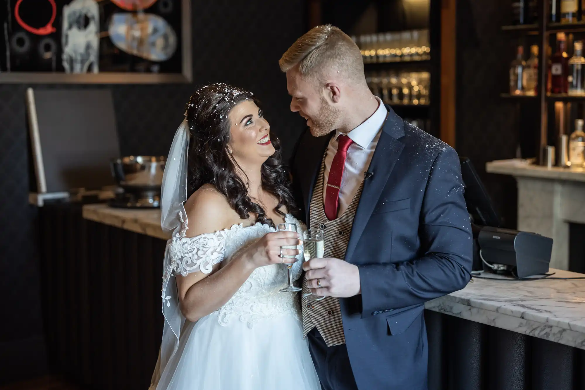 A bride and groom, dressed in a wedding gown and suit, smile and look into each other's eyes while holding champagne glasses in a bar.