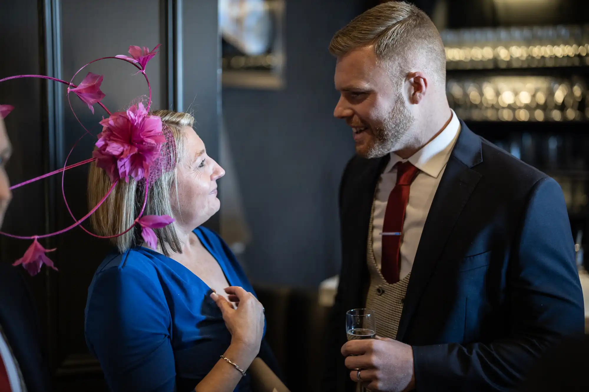 A woman wearing a blue dress and pink fascinator talks with a man in a dark suit and red tie at an indoor event. Both appear to be engaged in conversation, with the woman gesturing towards herself.