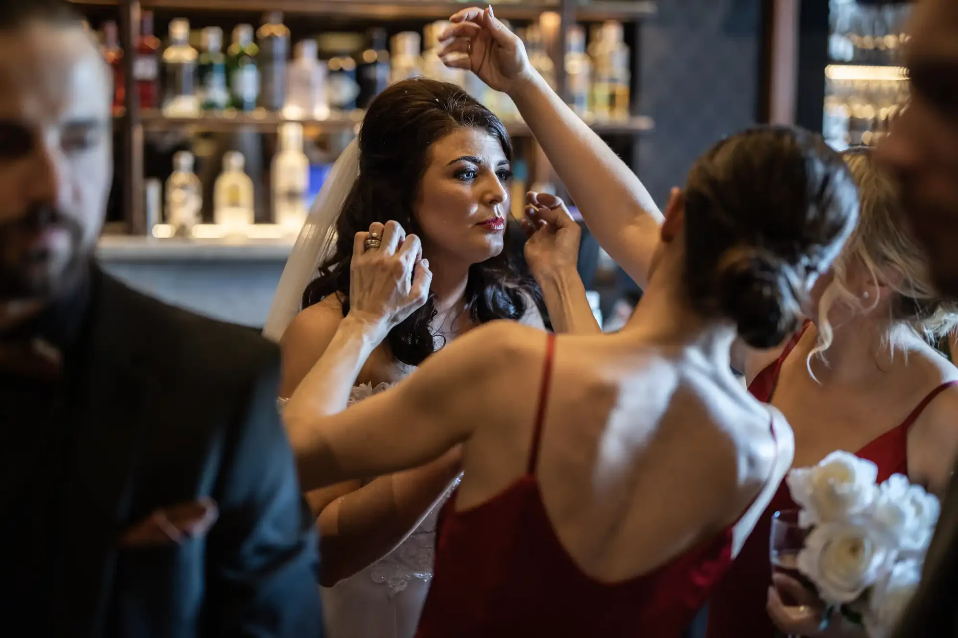 A bride gets assistance with her veil from a bridesmaid in a red dress, surrounded by other members of the wedding party in a bar setting.