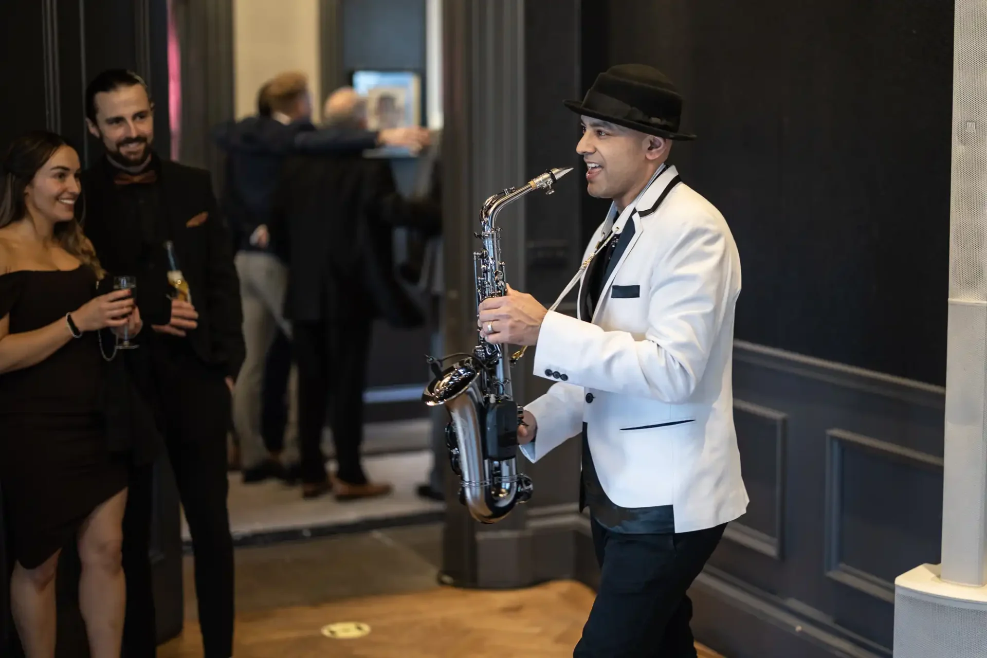 A man in a white jacket and hat plays the saxophone at an indoor event, with a couple holding drinks in the background.