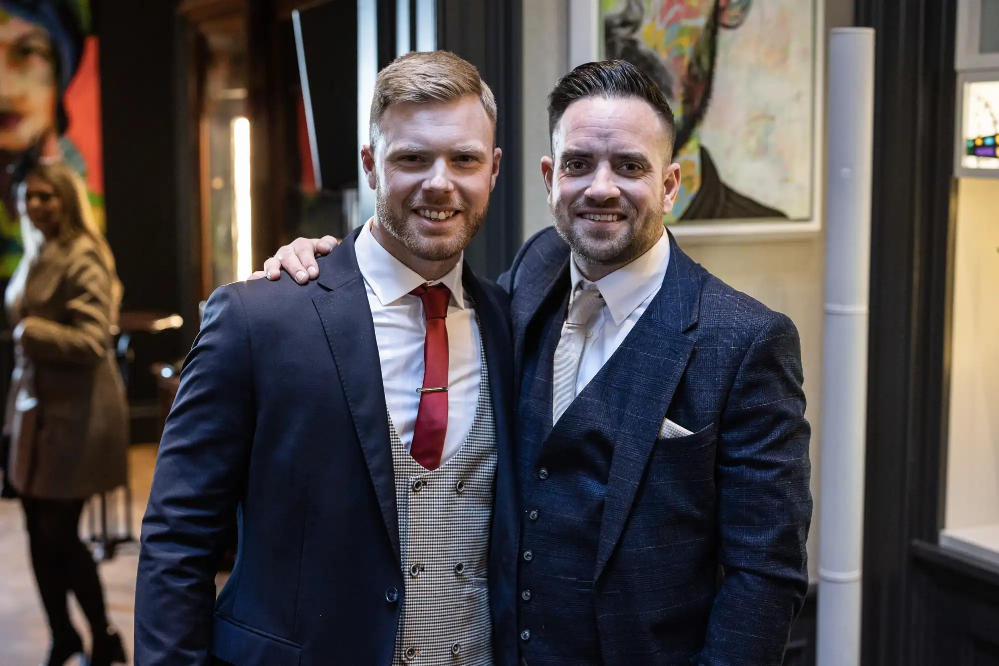 Two men dressed in suits are smiling and posing together indoors. One has a red tie, and the other has a blue tie. A person is blurred in the background.
