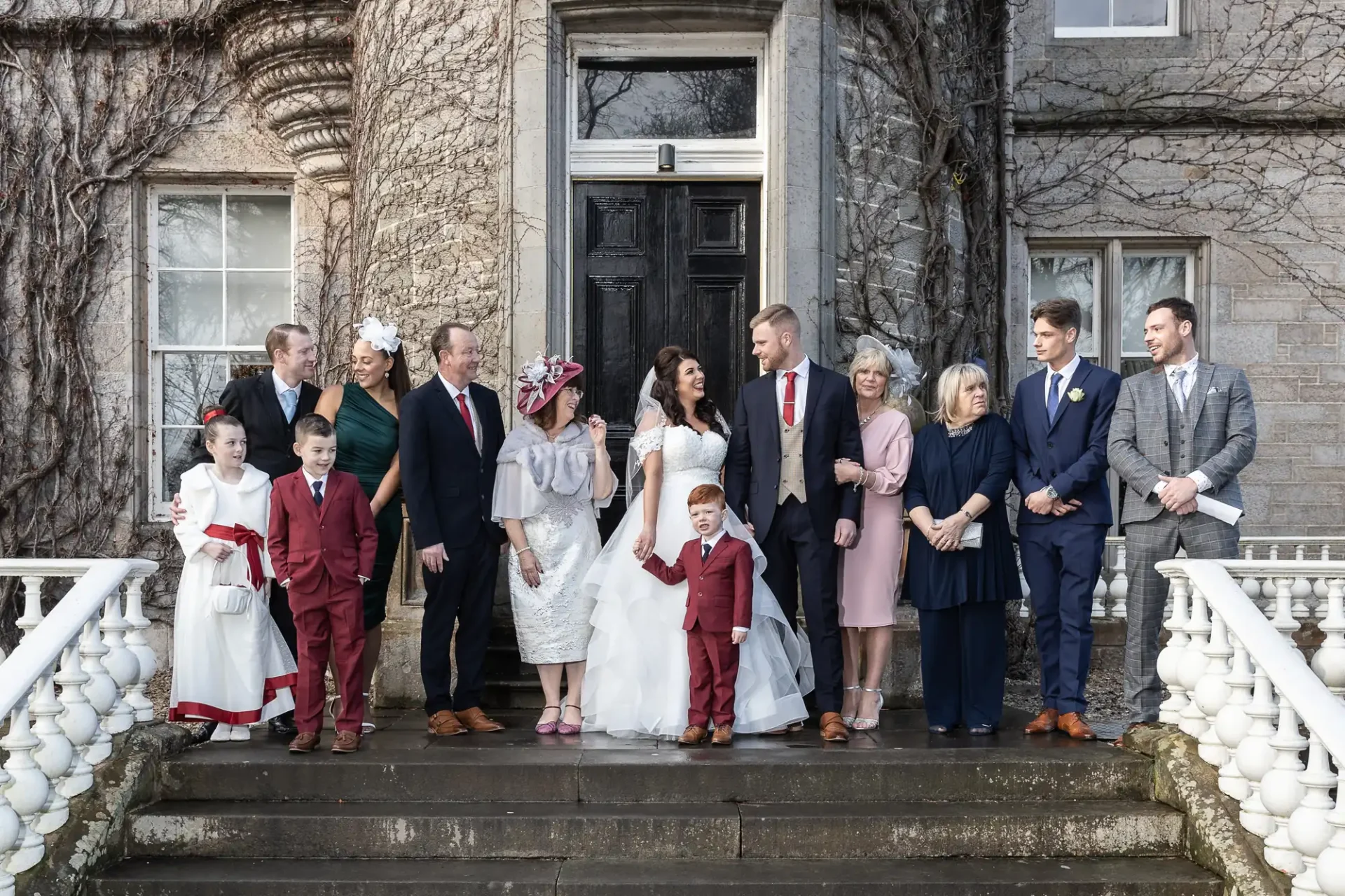 A large family gathers on steps outside a stone building, dressed formally for a wedding with smiles and varied interactions.