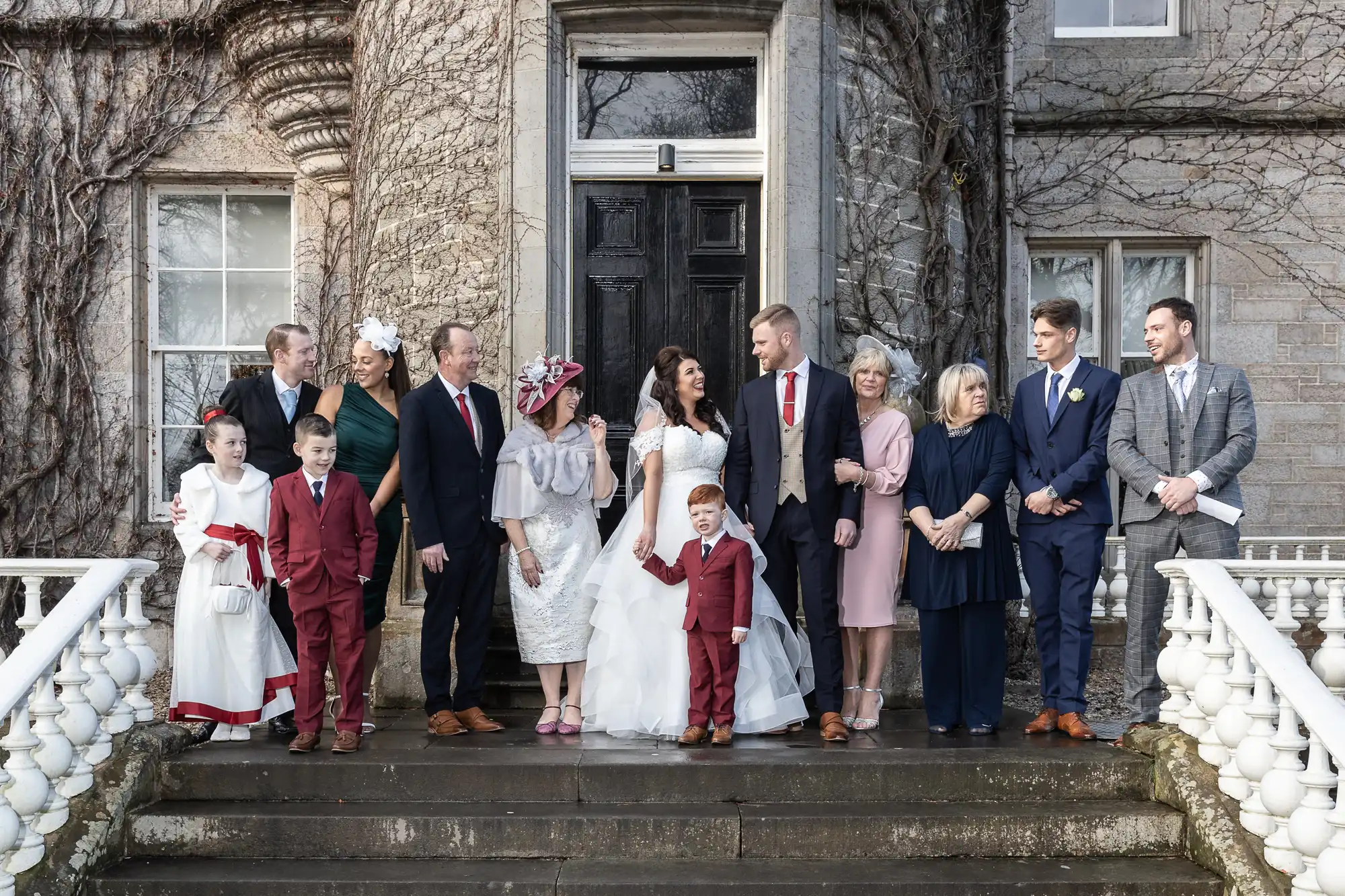 A wedding group photo featuring the bride, groom, and their family members dressed in formal attire, standing on the steps of an old stone building with vines.