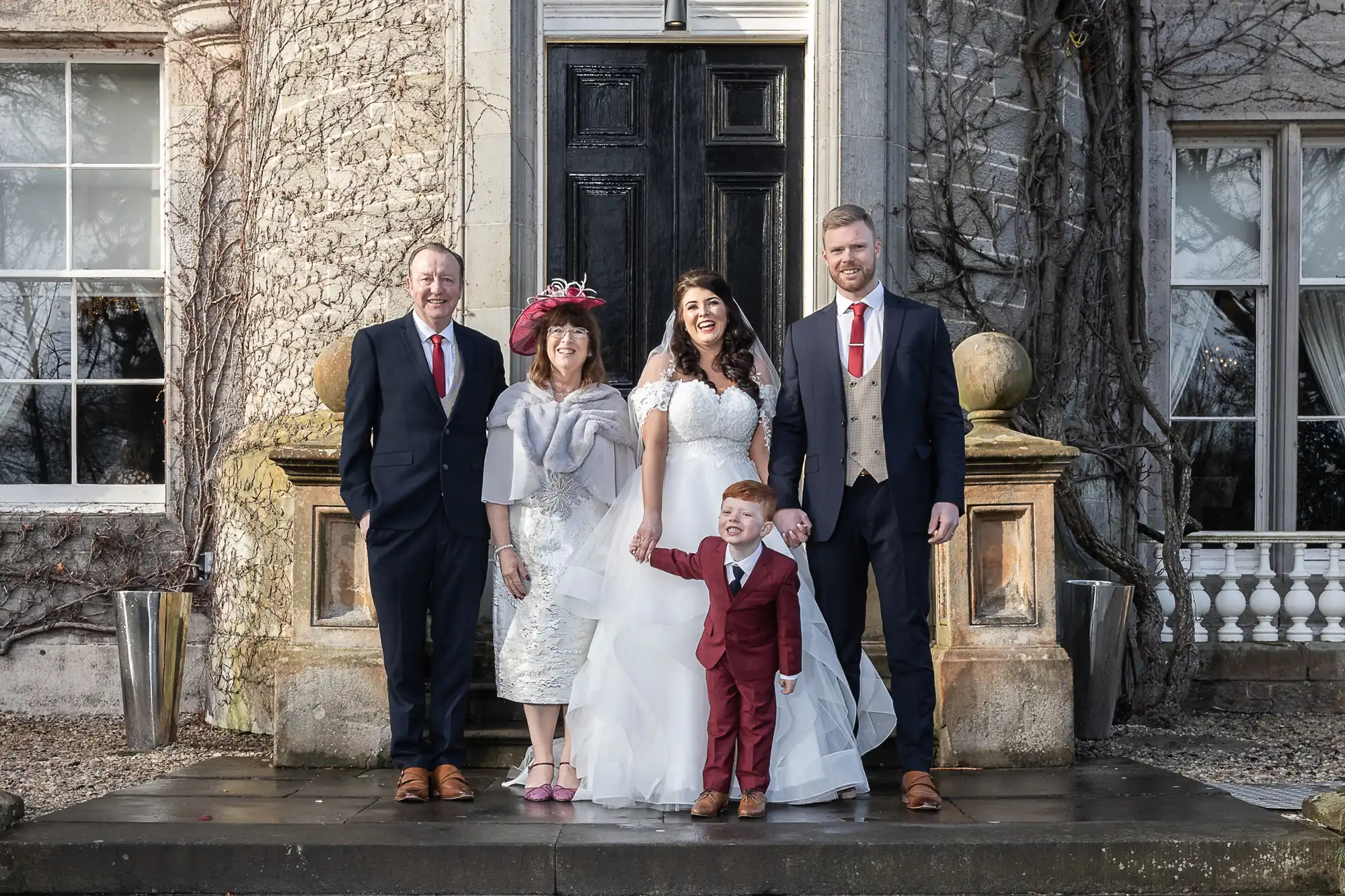 A family of five dressed in formal attire poses for a photo outside a large, old stone building with a black door. The child is wearing a red suit, and the adults are dressed in coordinated outfits.