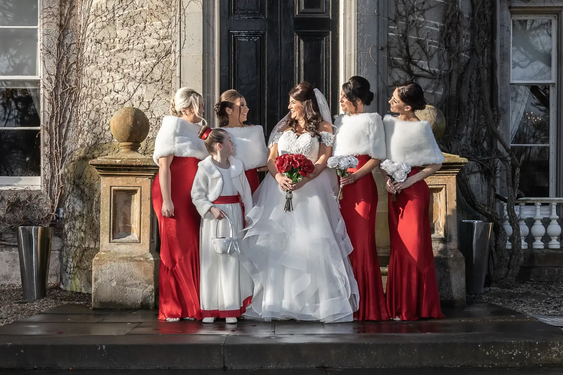 Bride in a white gown surrounded by bridesmaids in red dresses and white shawls, standing in front of a stone building.