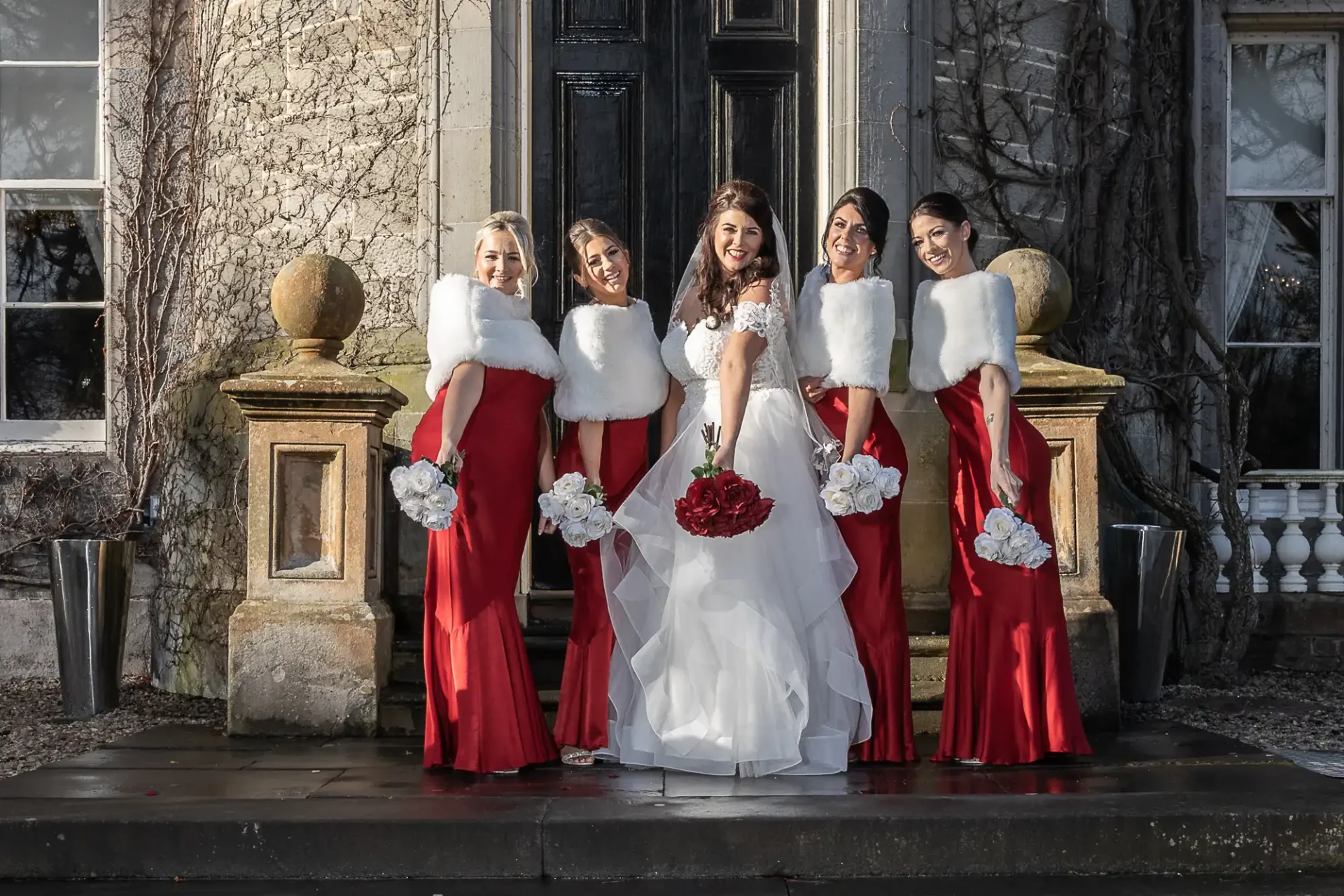 A bride and four bridesmaids posing in front of a building, wearing red dresses with white stoles and holding bouquets.