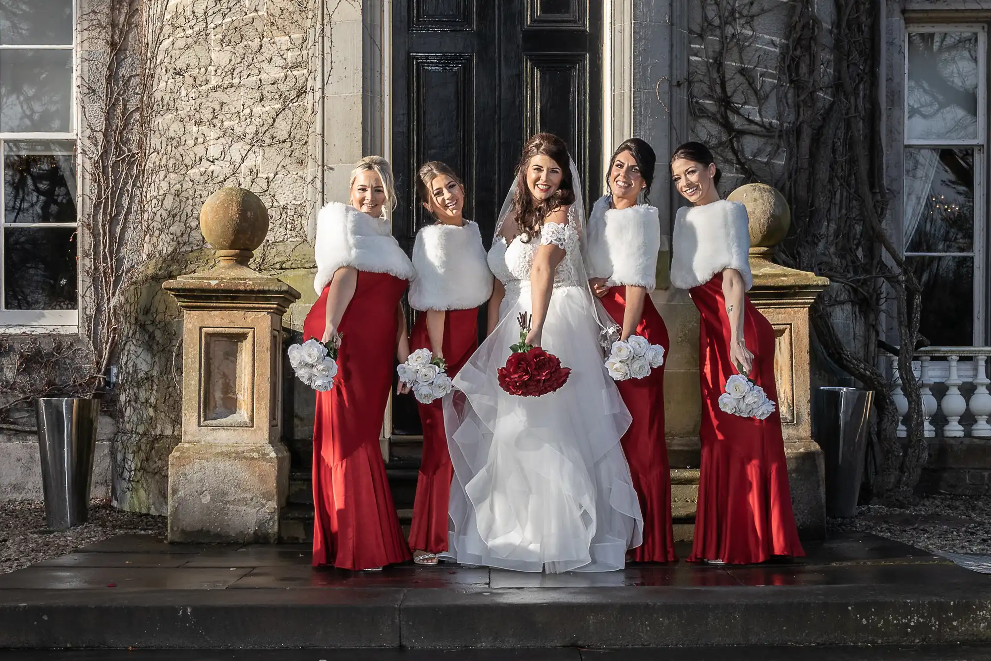 A bride in a white gown and floral bouquet stands with four bridesmaids in red dresses and white fur stoles, posing together outdoors in front of a large door.