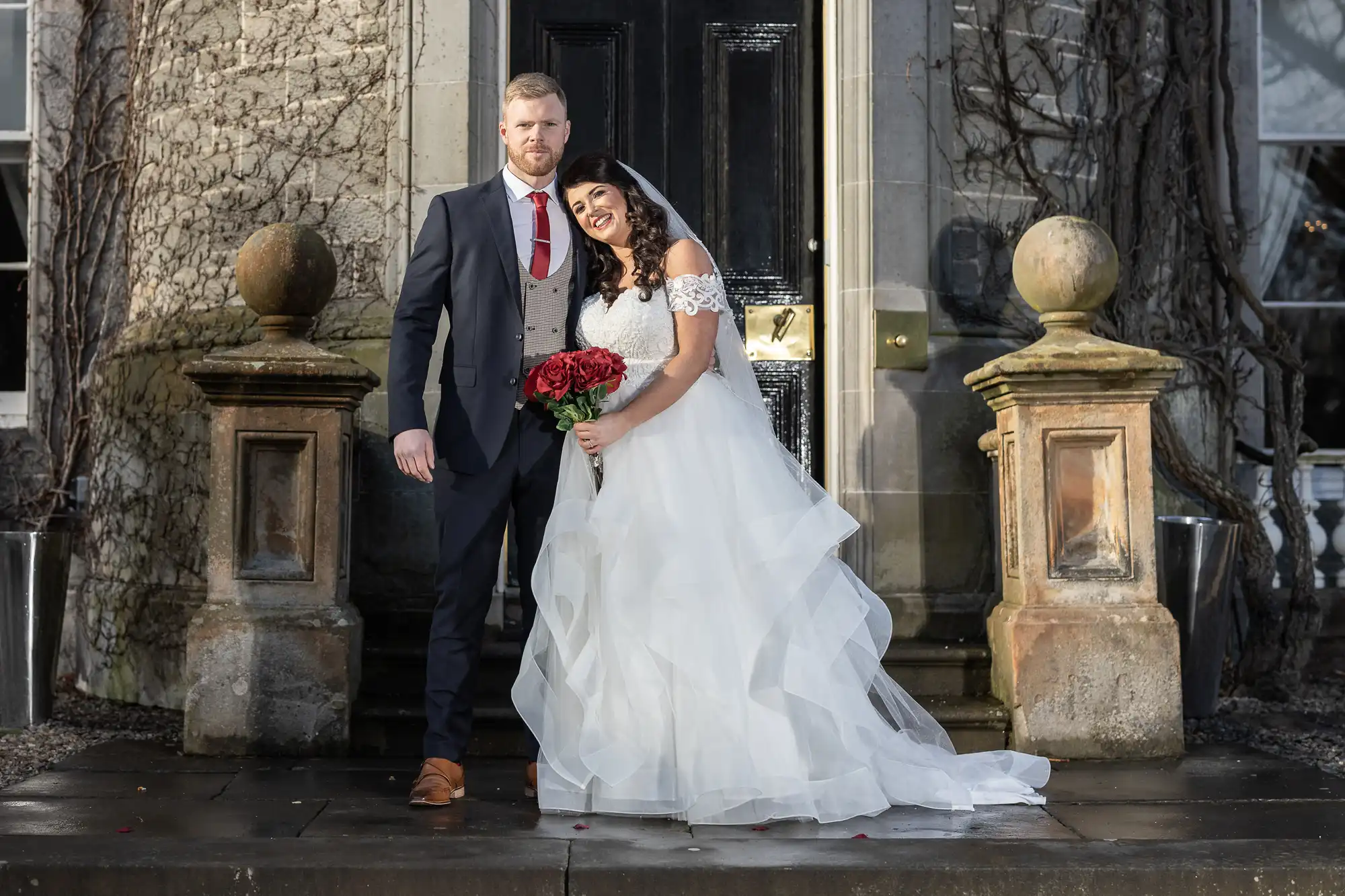 A bride in a white wedding dress holds red roses and stands next to a groom in a dark suit in front of a building entrance with ornate columns.
