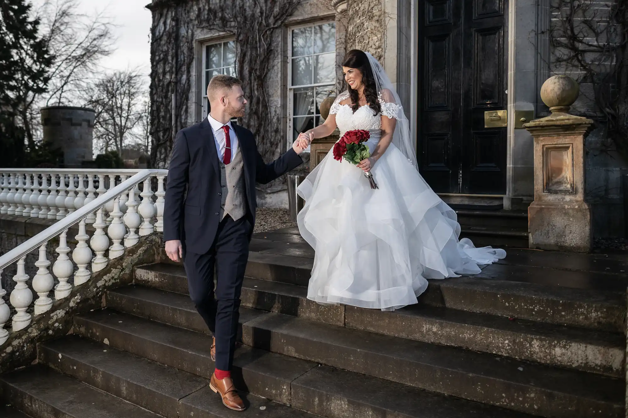 A couple descends the steps of a grand stone building. The man is dressed in a suit, and the woman wears a white wedding dress, holding a bouquet of red flowers.