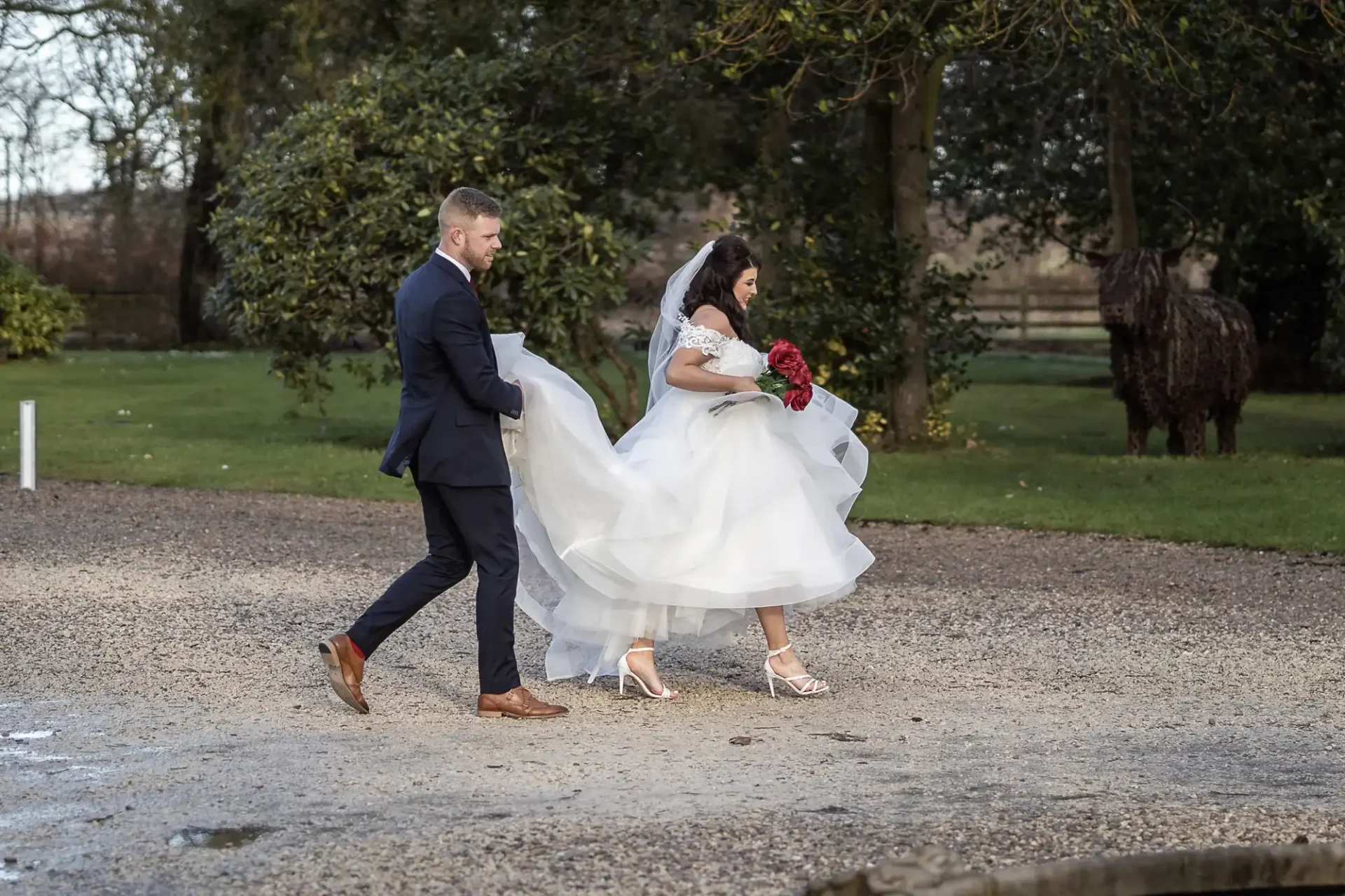 A bride and groom walk hand in hand across a gravel path, the bride holding up her white dress, both dressed formally.