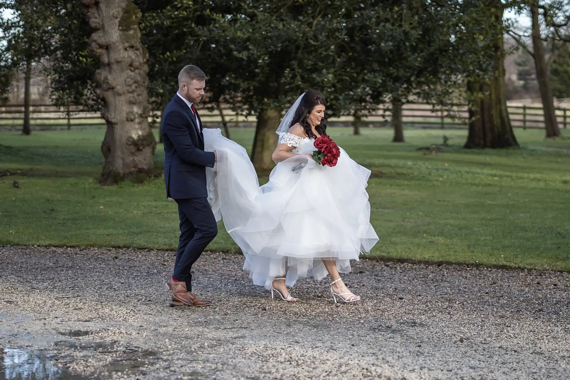 A bride and groom walk outside on a gravel path. The groom holds up the bride's white dress as she holds a bouquet of red flowers. Trees and fencing are visible in the background.