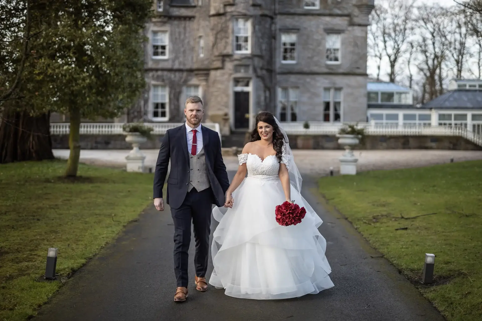 A bride and groom walking hand in hand in front of a historic stone castle, the bride holding a red bouquet.