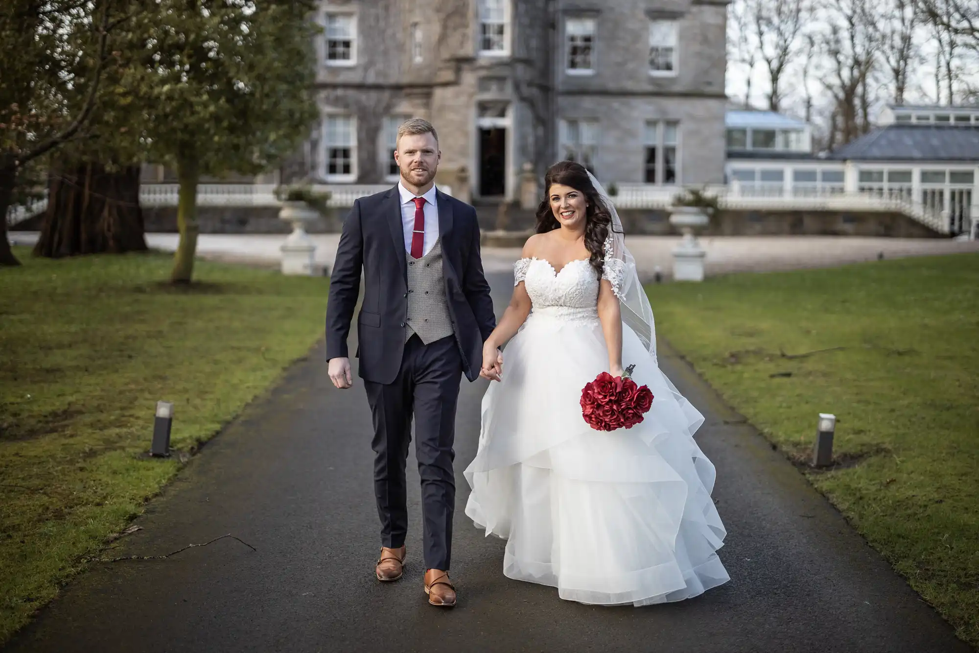 Carlowrie Castle wedding photos: A bride and groom walk hand-in-hand on a path in front of a large stone building. The bride is in a white dress holding red flowers; the groom is in a dark suit with a gray vest.