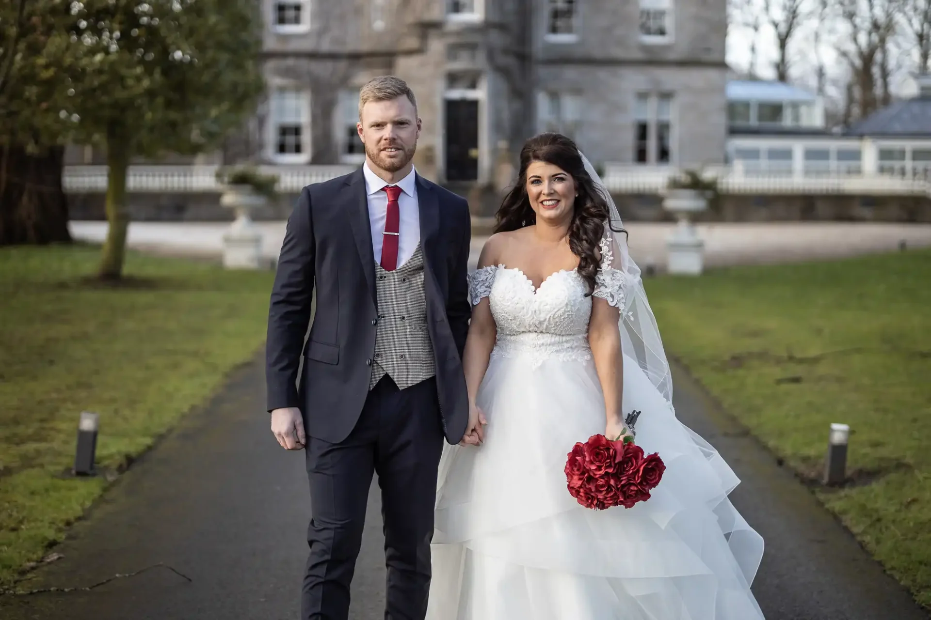 A bride in a white gown and a groom in a gray suit with a red tie walk hand in hand in front of a stone manor house.