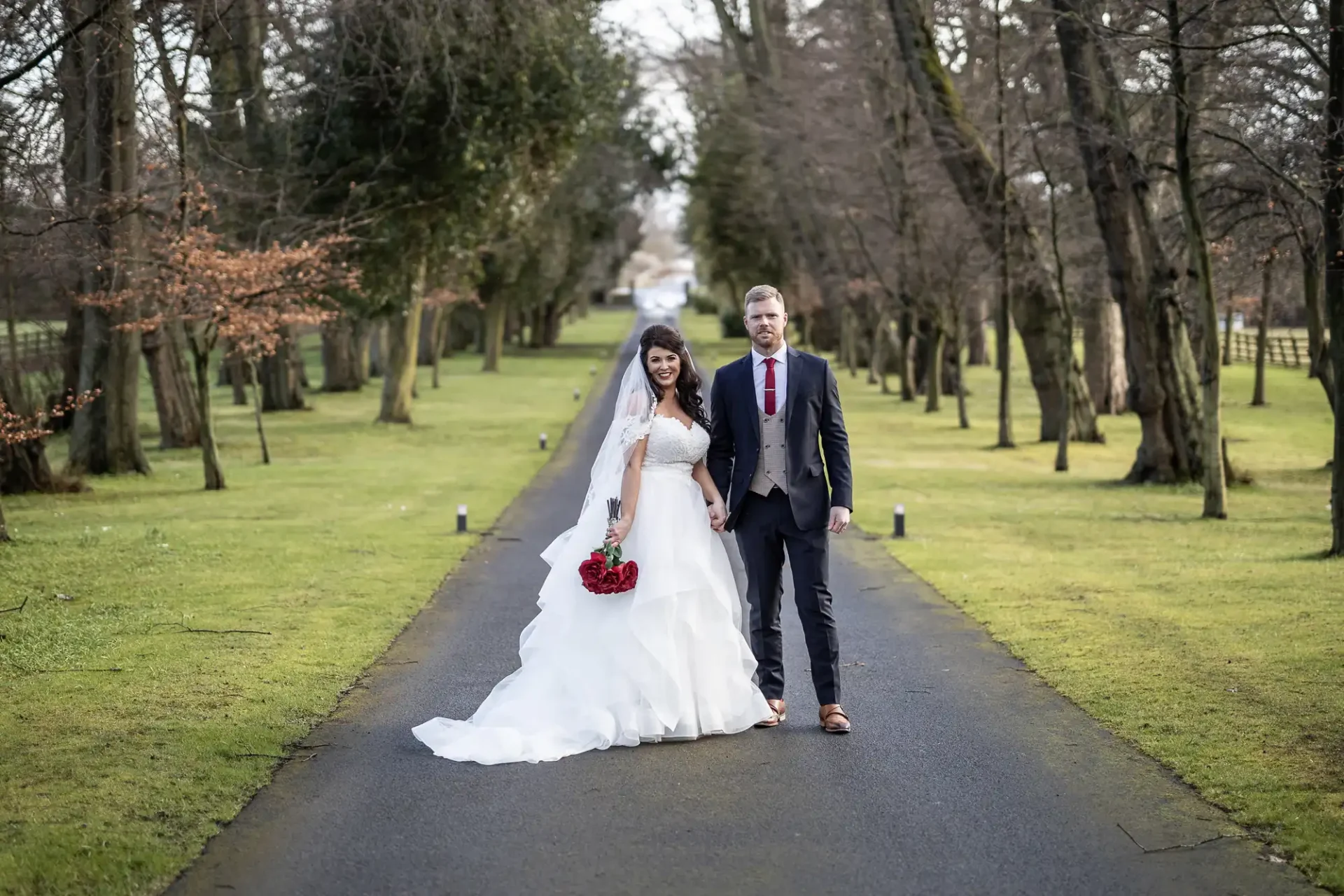 A bride and groom holding hands, walking down a tree-lined pathway, the bride in a white dress and the groom in a suit with red tie.