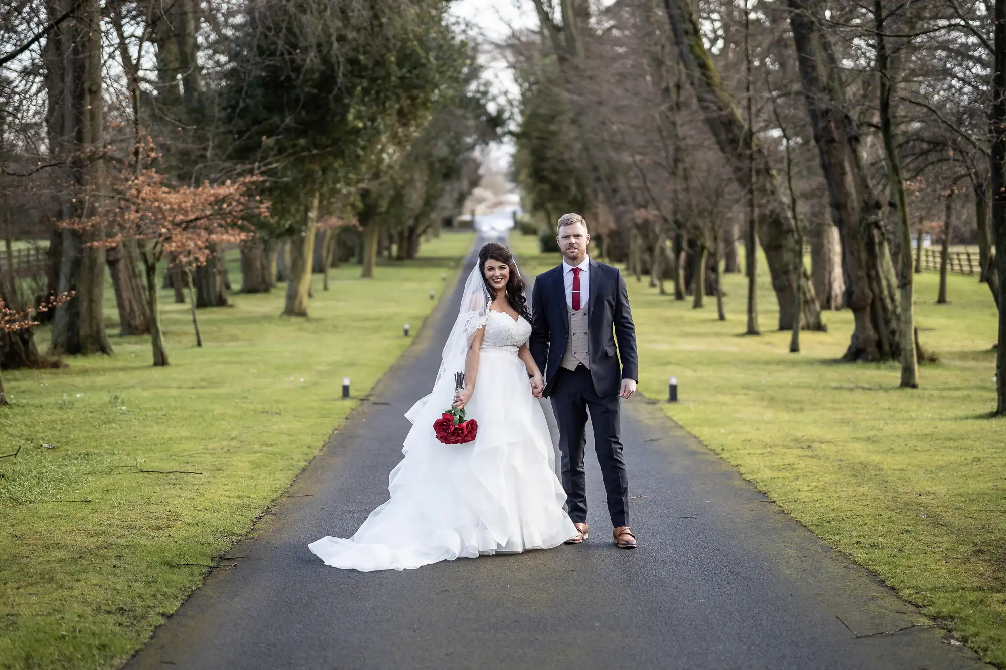 A bride and groom, dressed formally, stand hand in hand on a tree-lined pathway. The bride holds a red bouquet, and both are looking towards the camera.
