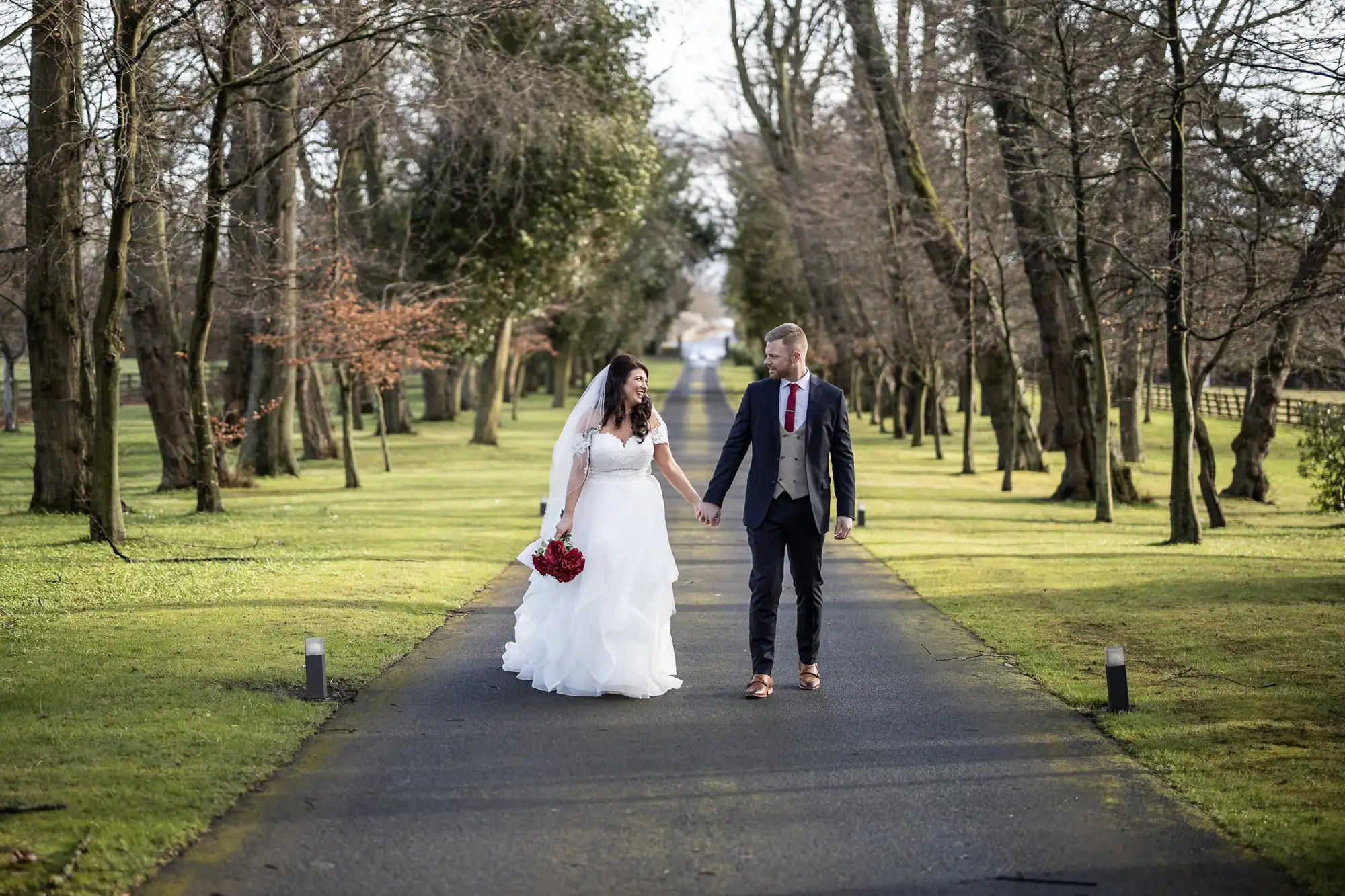 A bride in a white gown and a groom in a suit walk hand in hand on a tree-lined path, surrounded by bare trees and green grass. The bride carries a bouquet of red flowers.