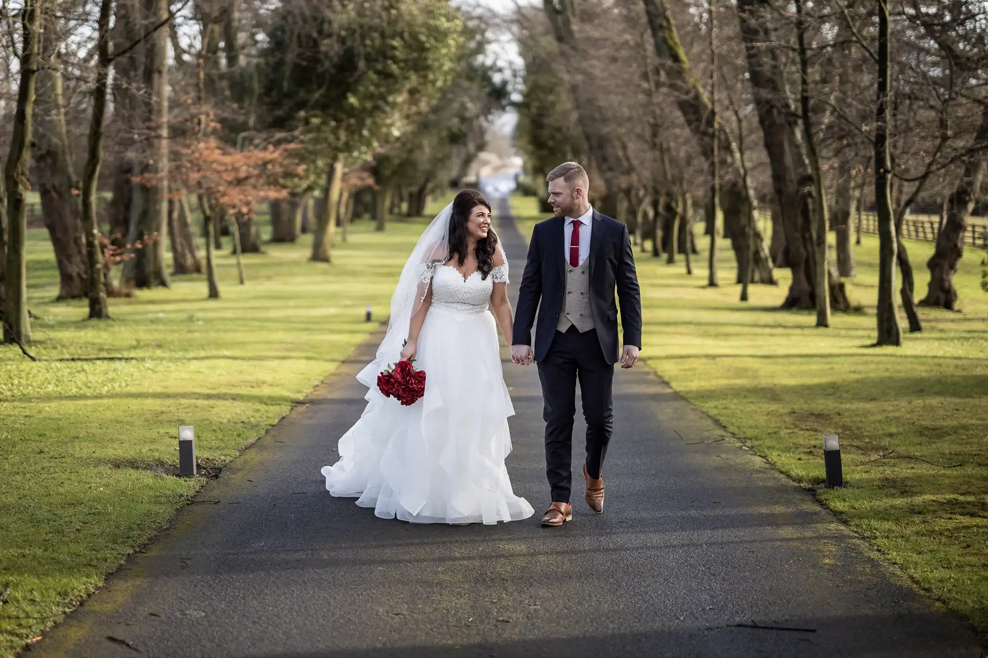 A couple in wedding attire walks hand in hand down a tree-lined path. The bride holds a red bouquet and wears a white dress with a veil, while the groom wears a suit with a red tie.