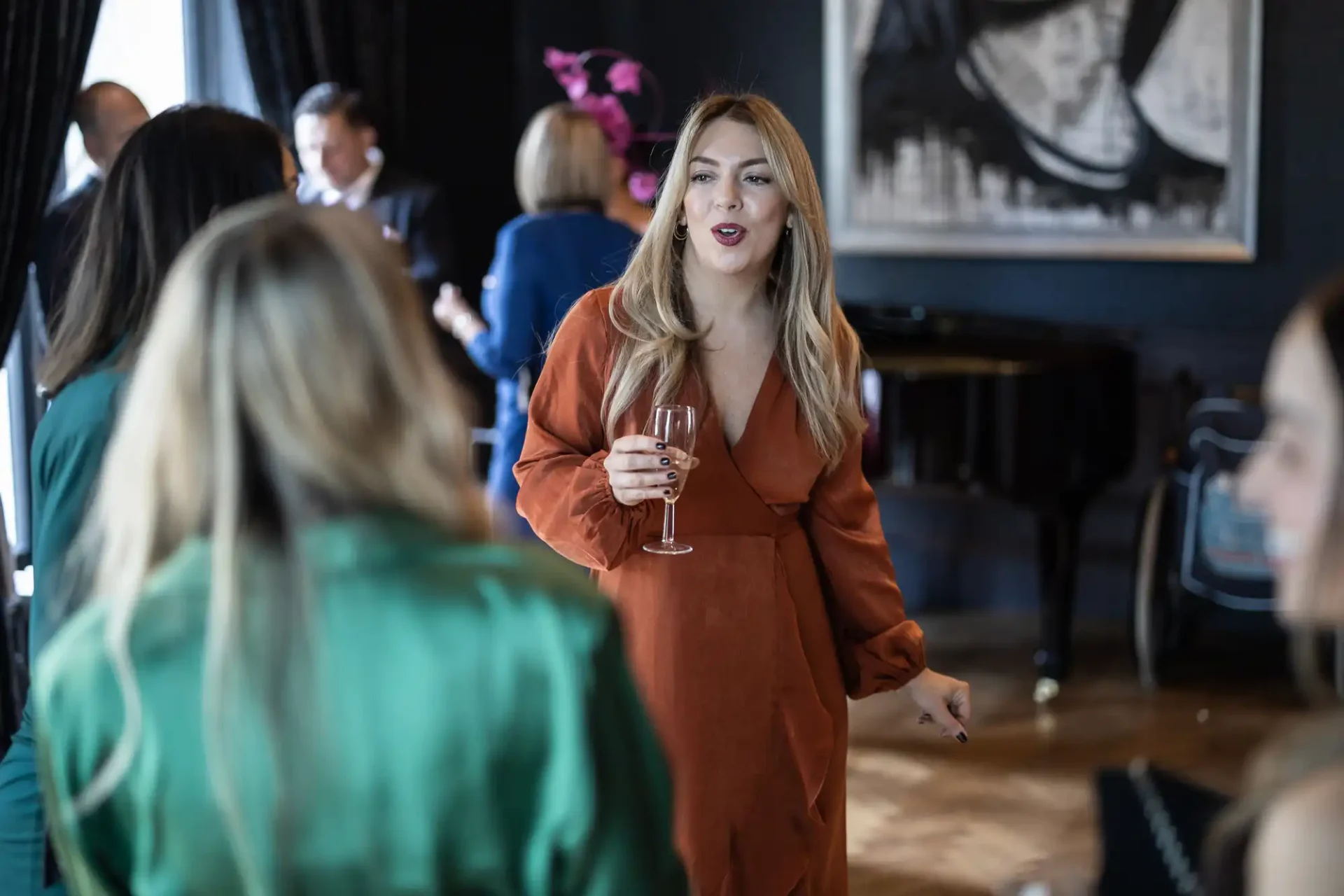 Woman in an orange dress holding a wine glass, conversing with others at a social event indoors.