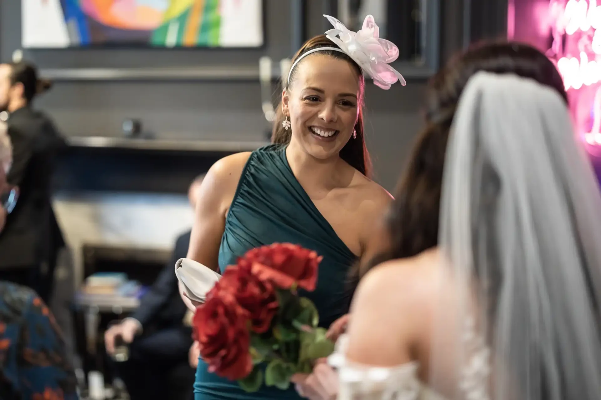 A bridesmaid in a teal dress and fascinator smiles as she holds a bouquet of red roses, talking to a bride in a white dress.