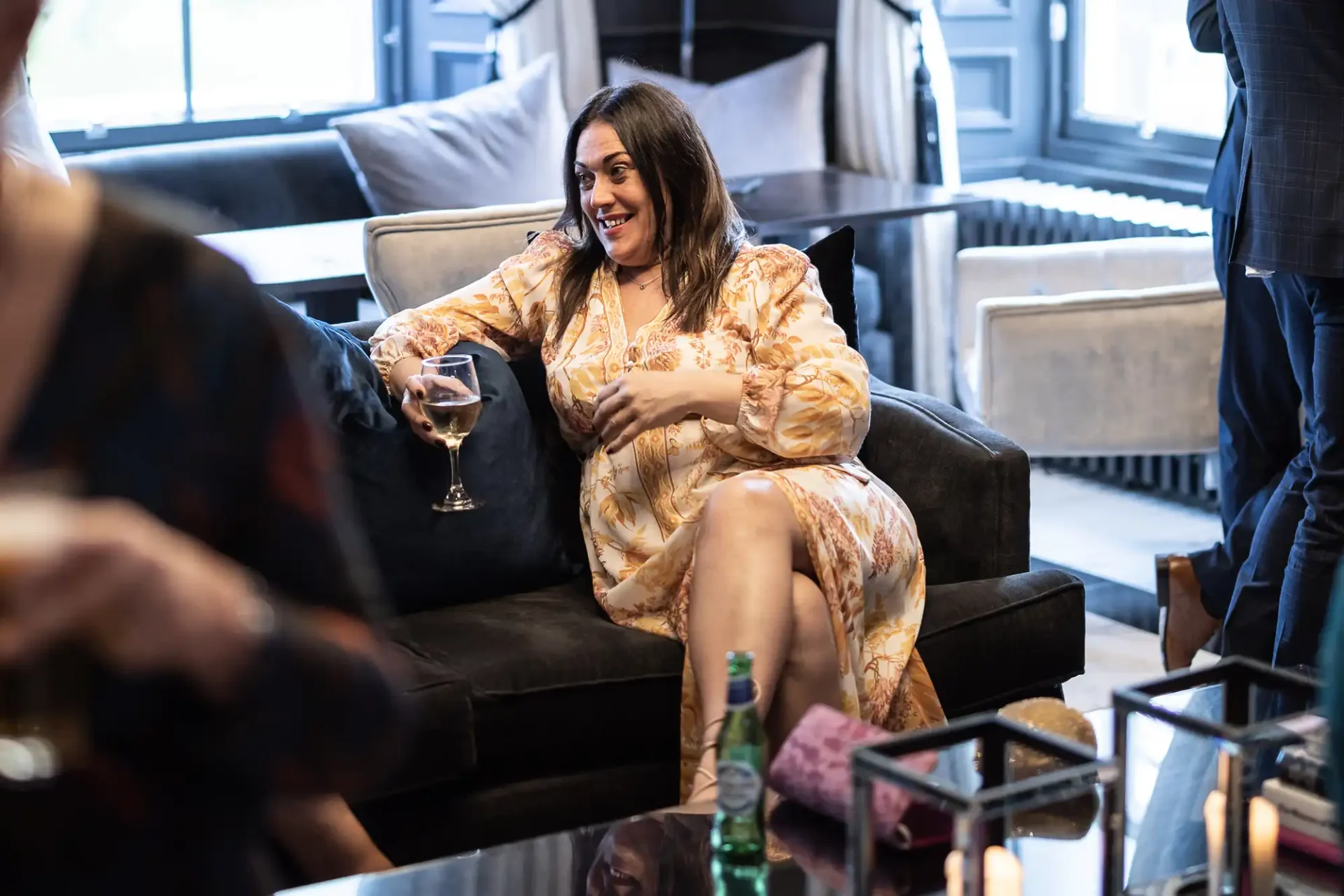 A woman in a floral dress, laughing and holding a wine glass, sits on a black sofa in a stylish lounge.