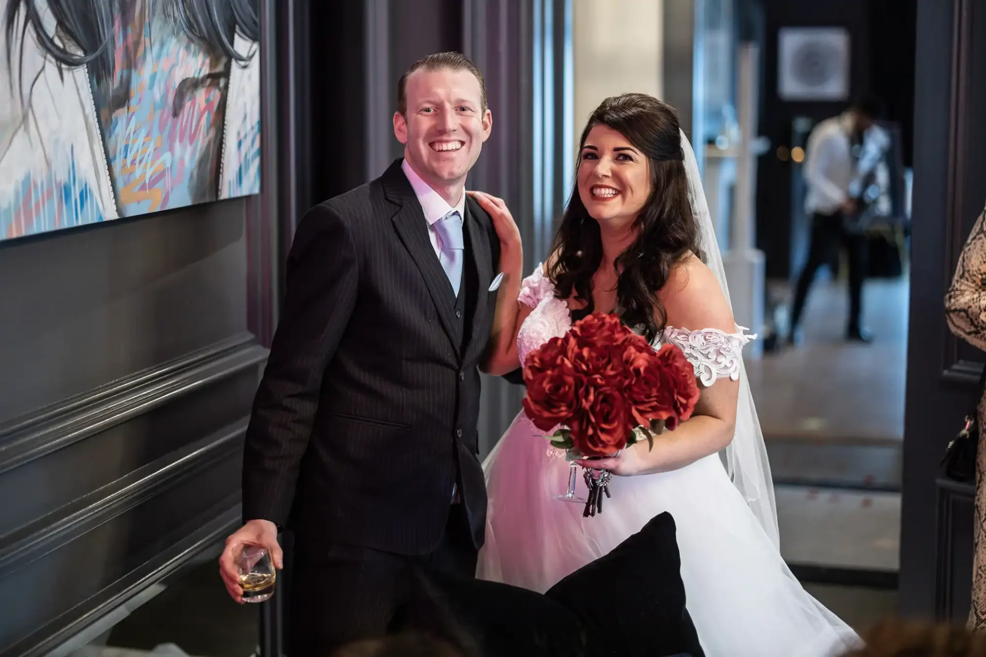 A newlywed couple smiling joyously; the groom in a black suit holds a drink, and the bride in an off-shoulder gown clutches a bouquet of red roses.