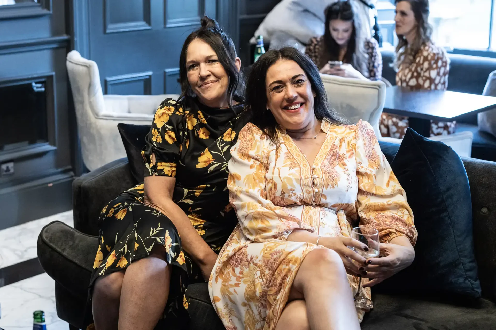 Two women smiling and sitting on a couch in a lounge, one wearing a black floral dress and the other in a cream floral dress.