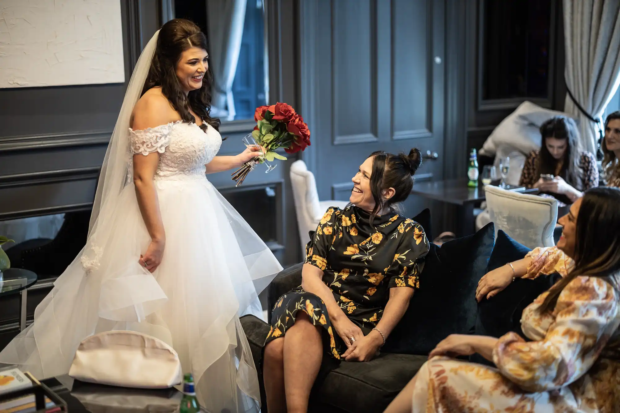 A bride in a white dress and veil holds a bouquet of red roses while talking to two seated women. Others are visible in the background, including a woman using a smartphone.