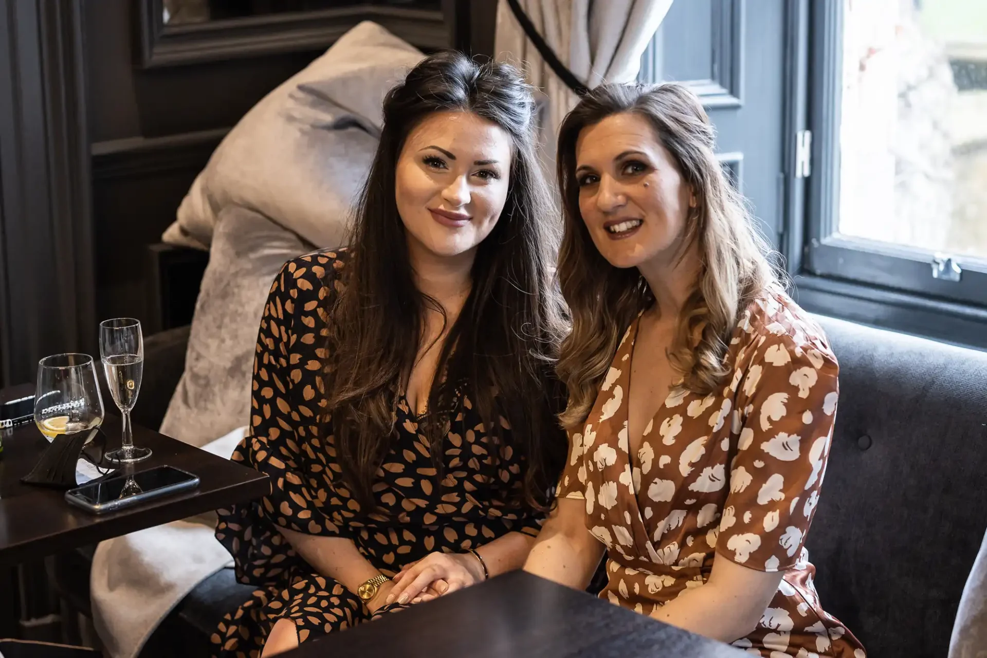 Two women in patterned dresses smiling at the camera while sitting with glasses of champagne in a cozy, dimly-lit lounge.