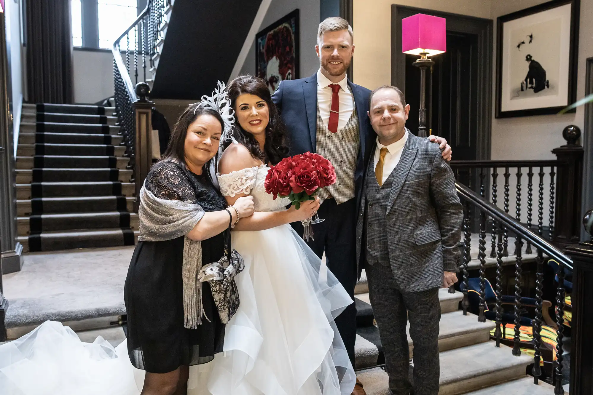 Four people pose for a wedding photo near a staircase. The bride holds a bouquet of red flowers and stands next to the groom, and two other guests flank them.