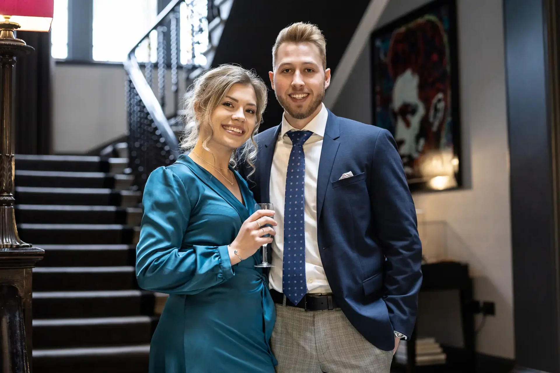 A smiling woman in a teal dress and a man in a suit holding a glass of champagne, standing together in an elegant hallway with a staircase.