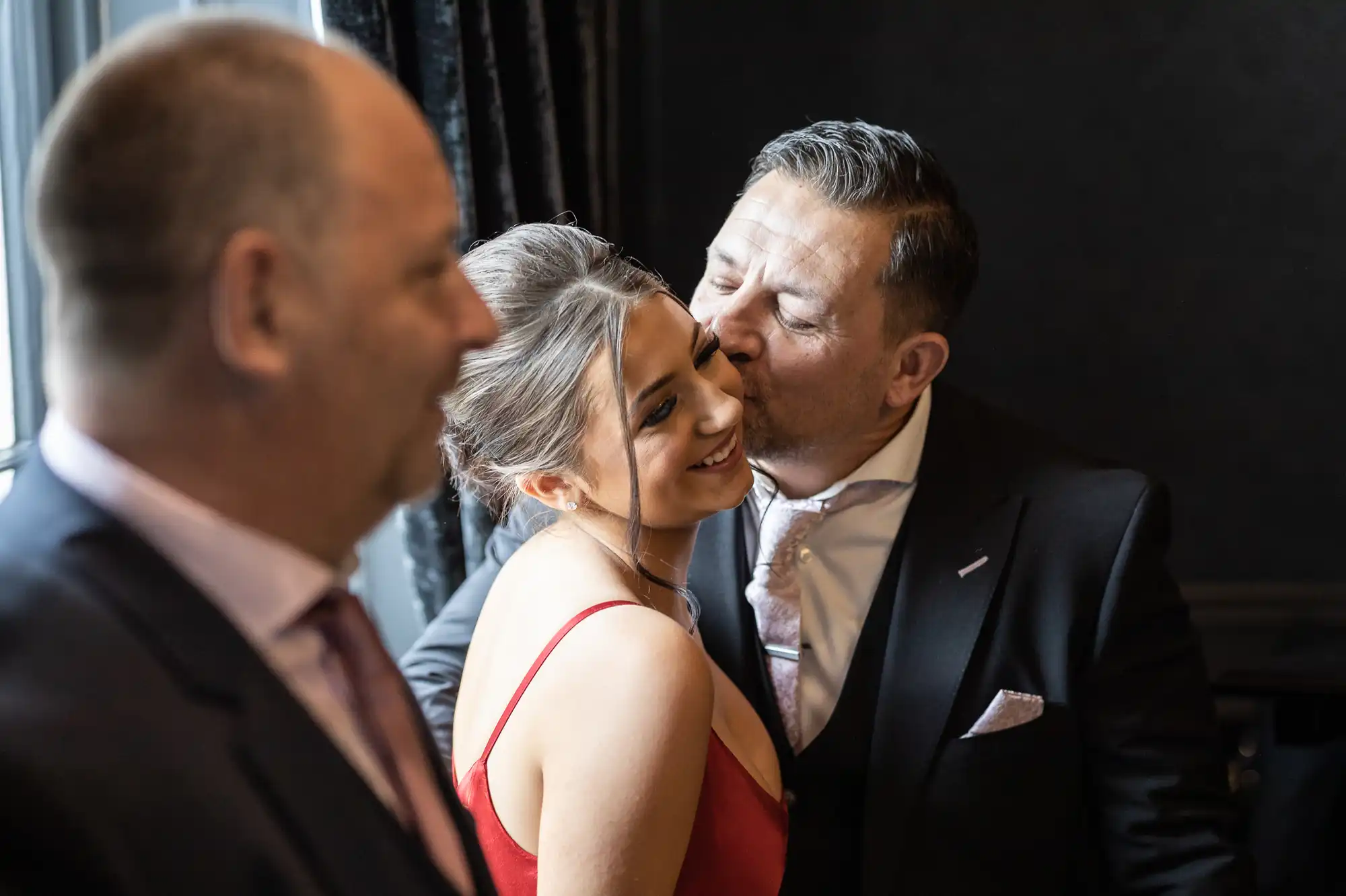 Three people in formal wear: a man on the left, a woman in a red dress smiling as a man in a suit kisses her cheek in an indoor setting.