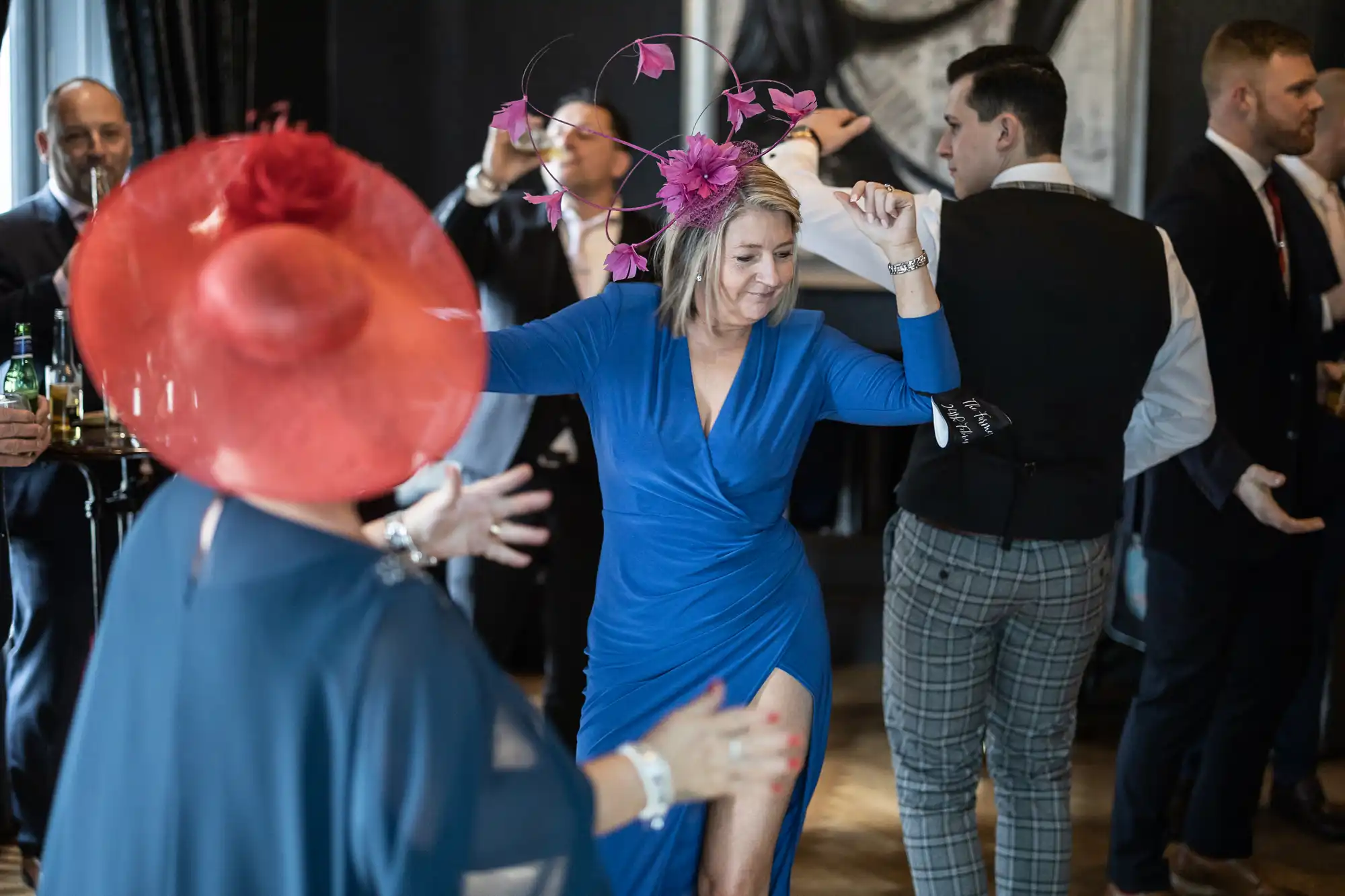 People dressed in formal attire are dancing at an indoor social event. A woman in a blue dress is prominently featured in the foreground, wearing a fascinator with pink flowers.