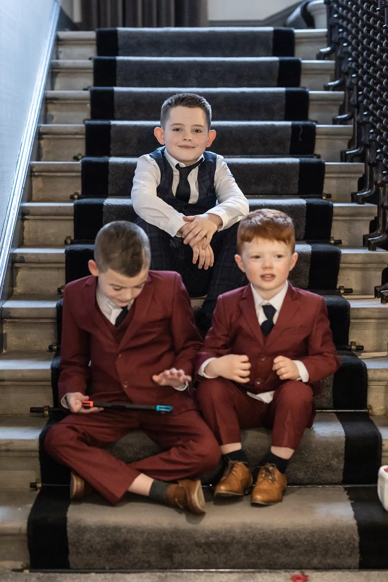 Three boys in formal wear sitting on a staircase; one in a gray vest and tie looking at the camera, two in maroon suits focused on a handheld device.