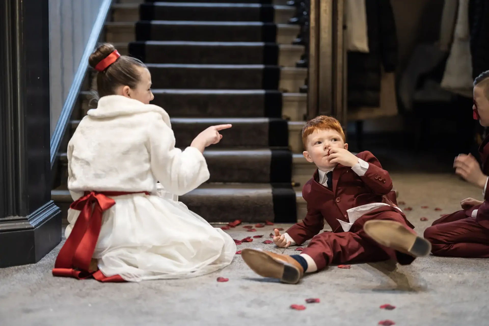 A young boy in a suit sits on the floor looking thoughtful while a girl in a white dress with a red sash points at him, both surrounded by rose petals.