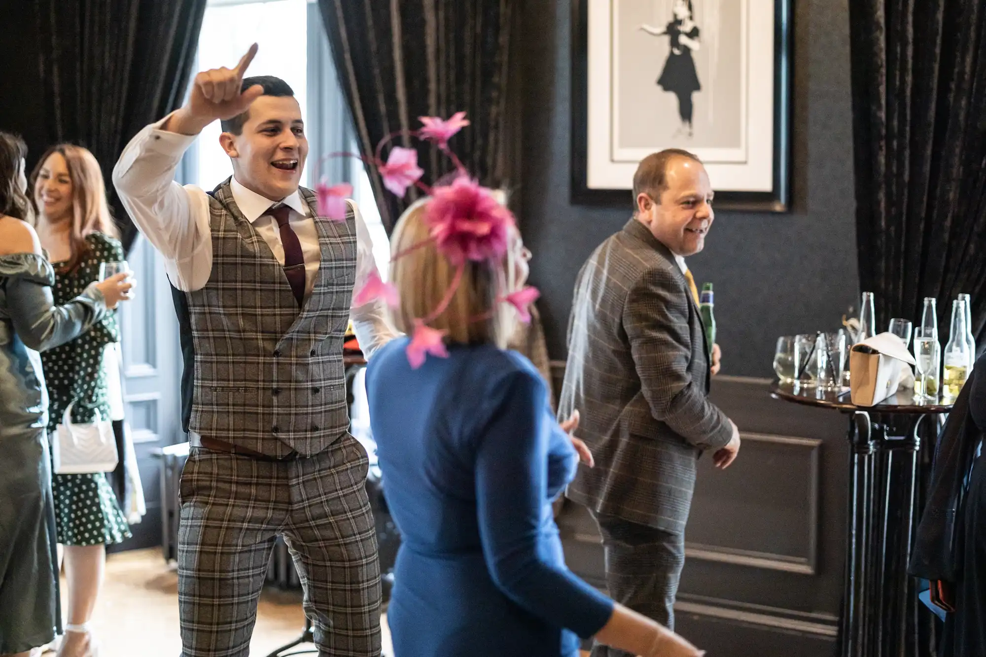 A group of people dressed formally is enjoying a social gathering. One man in a plaid suit is dancing energetically, while others are laughing and talking in the background.