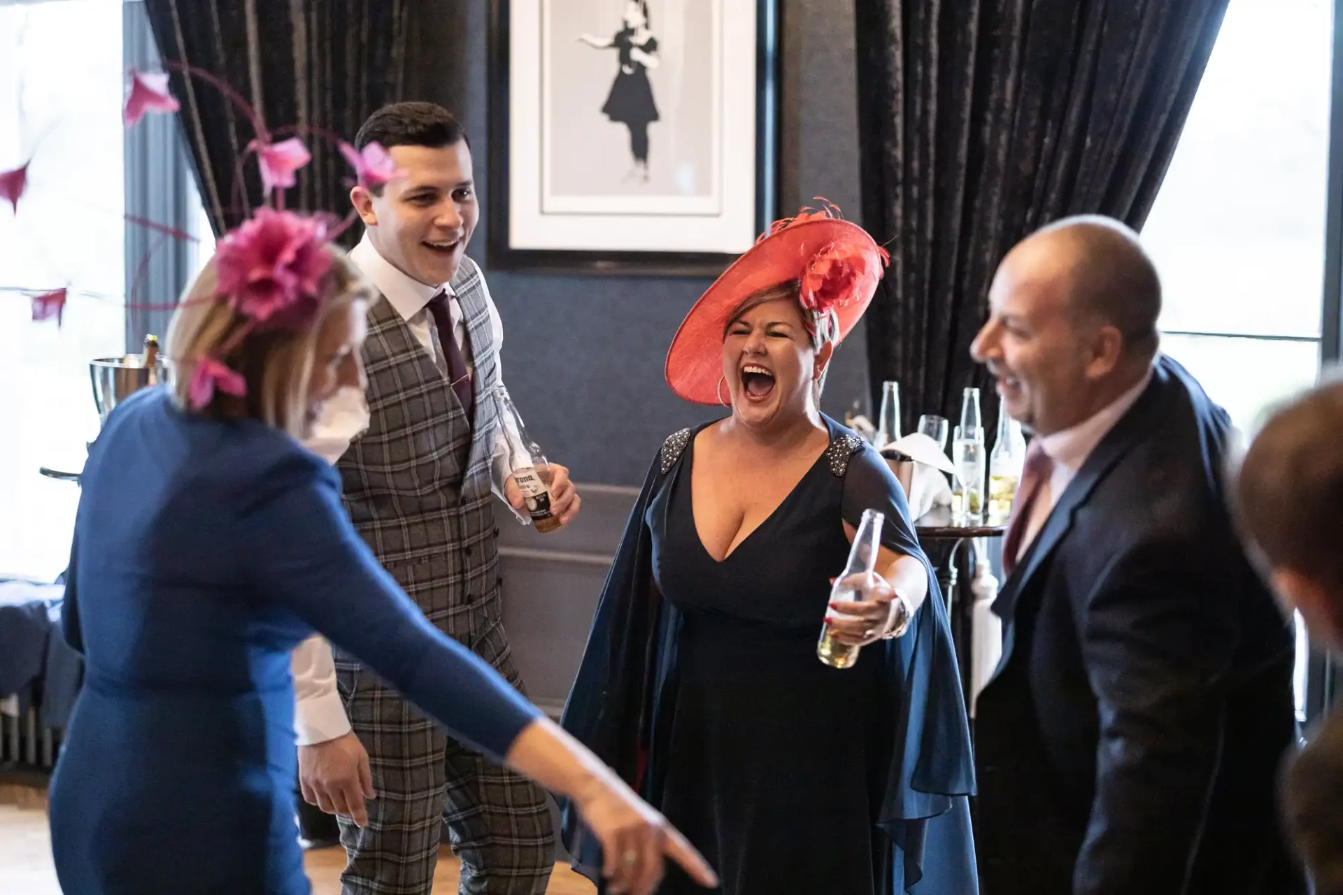 A woman in a red hat laughing joyously at a social gathering while holding a drink, surrounded by three other adults in a decorated room.
