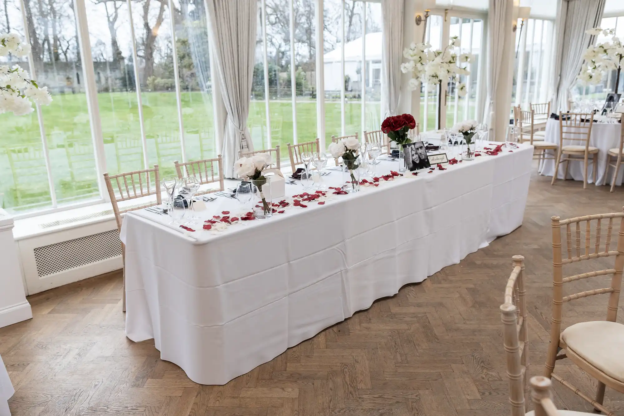 A long table with a white tablecloth, decorated with red and white flowers, and set with dining ware in a room with large windows and light wooden chairs.
