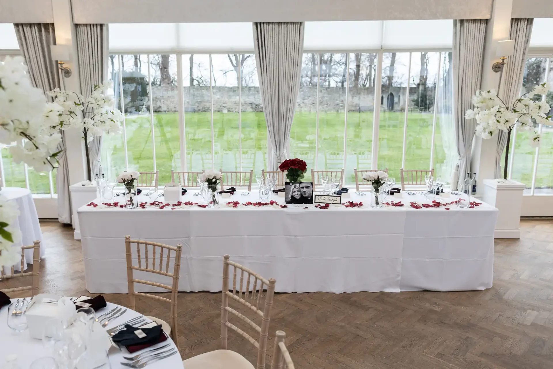 Elegant wedding reception table setting with white and red decorations, floral centerpieces, and framed photos, in a room with large windows overlooking a garden.