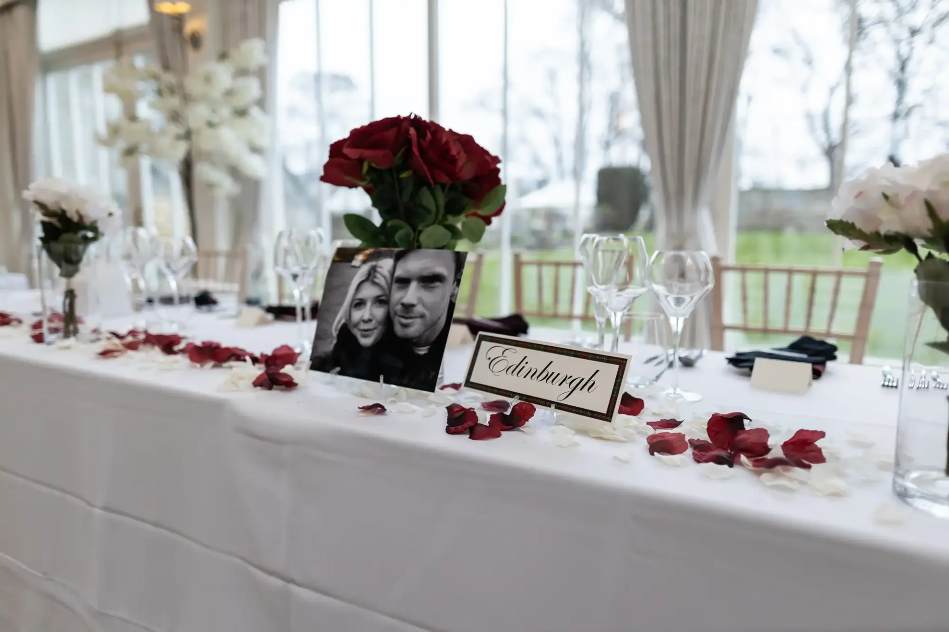 Decorated wedding reception table with a photo of a couple, red roses, scattered petals, and a table sign reading "edinburgh".