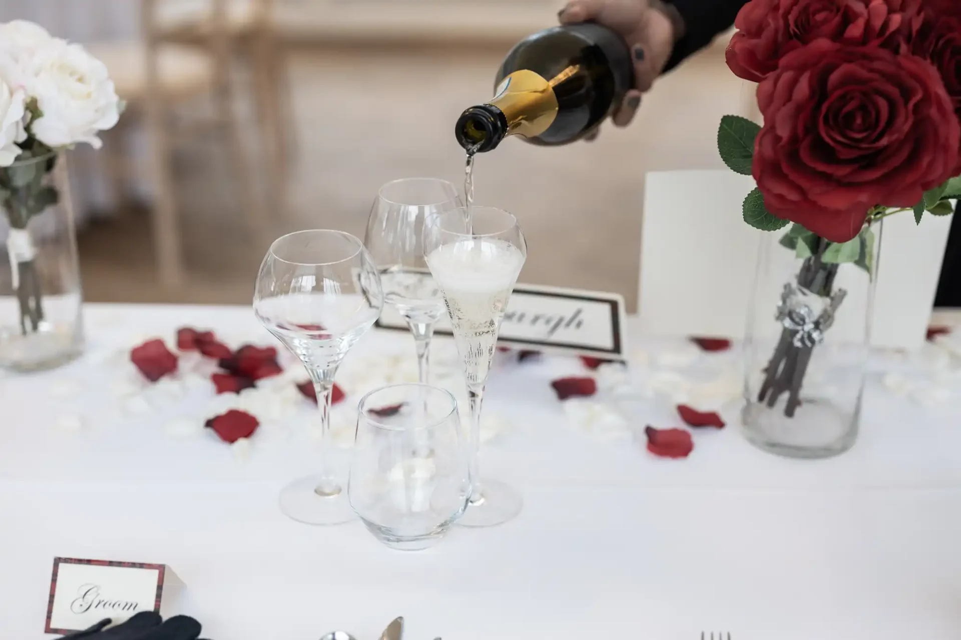 A hand pouring white wine into a tall glass at a formal dining table decorated with red roses and rose petals.