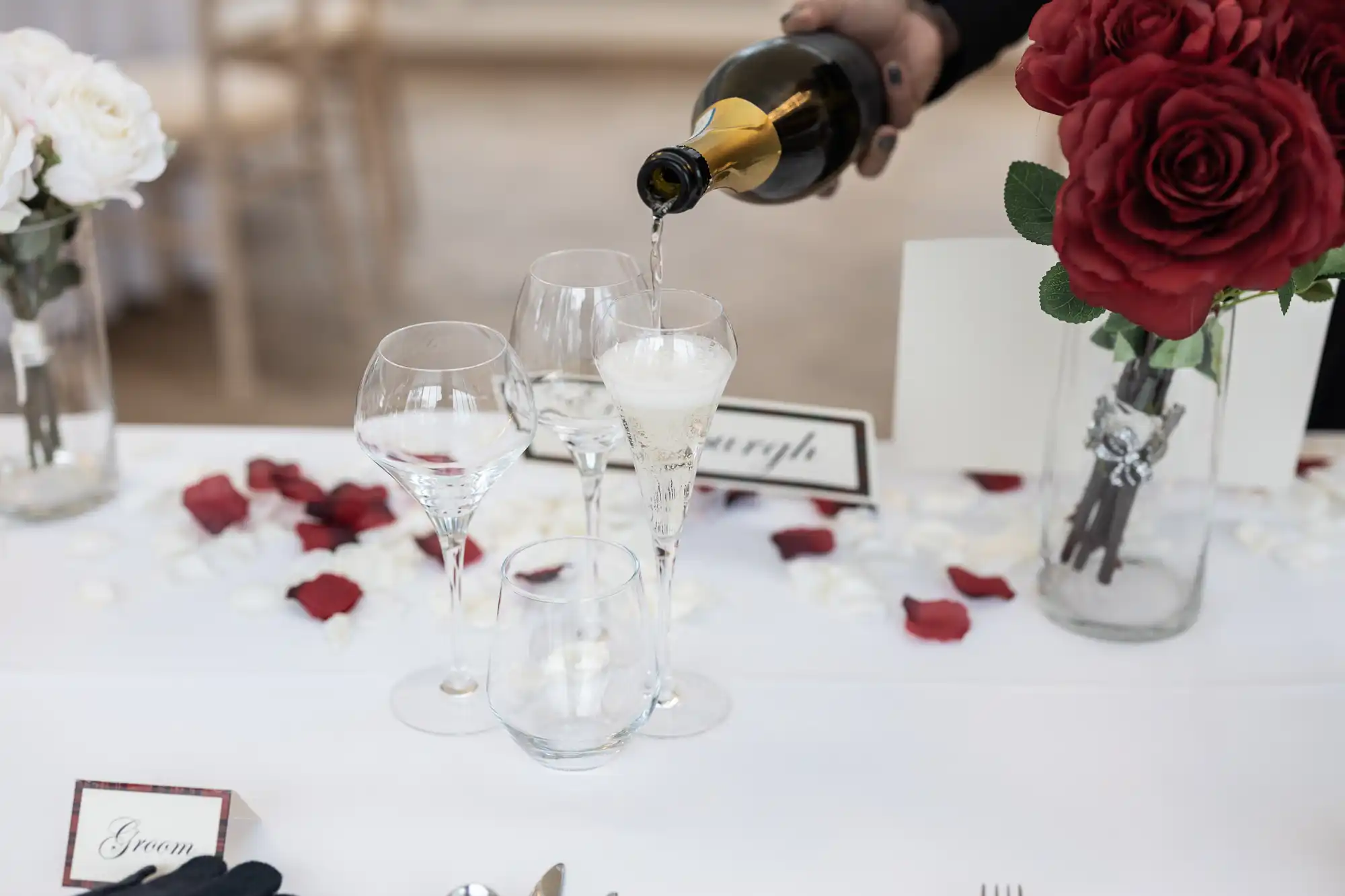 Hand pouring champagne into flute on elegantly decorated table with red, white roses, and scattered rose petals.