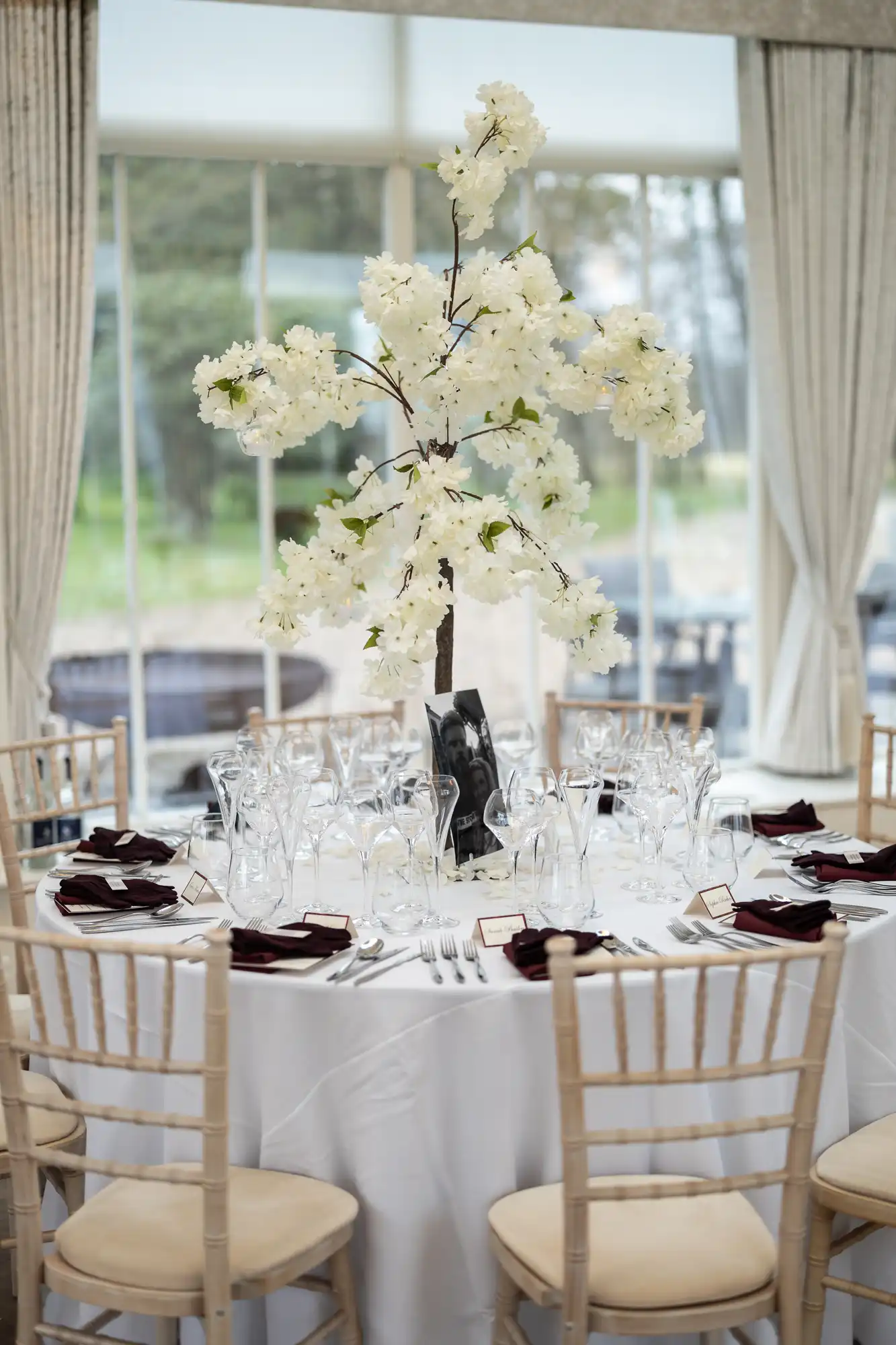 Elegant wedding table setup featuring white floral centerpiece, crystal glassware, and burgundy napkins in a venue with large windows and curtains.