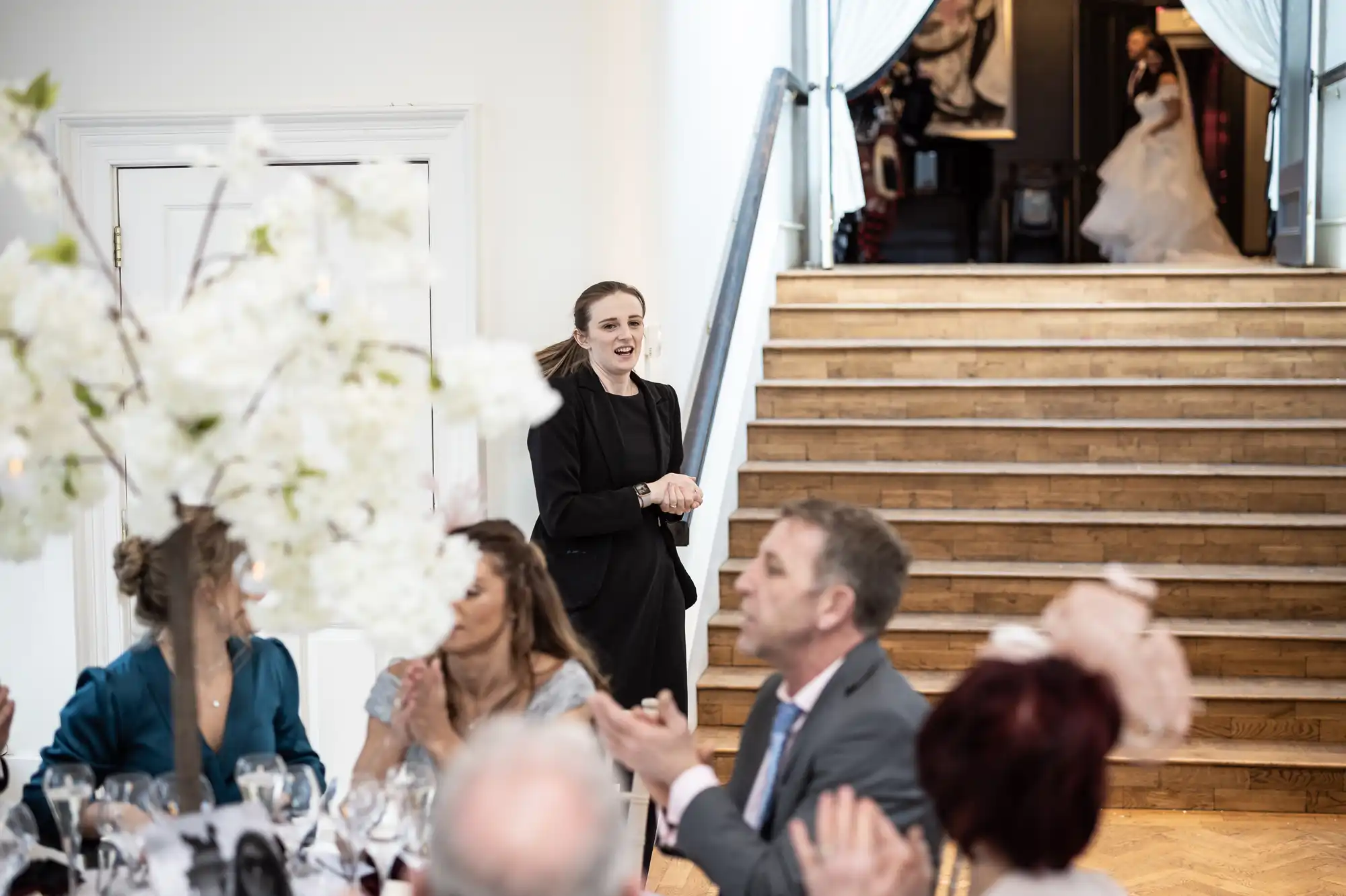 A woman in a black outfit stands near stairs, smiling, while guests at a table applaud in a bright room with floral decor. Another woman in a white dress is partially visible at the top of the stairs.