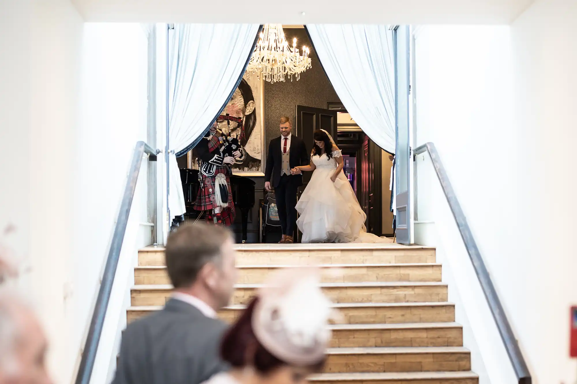 A bride in a white gown and a groom in a suit walk down steps, holding hands, with a chandelier and draped curtains in the background. Guests watch them descend.