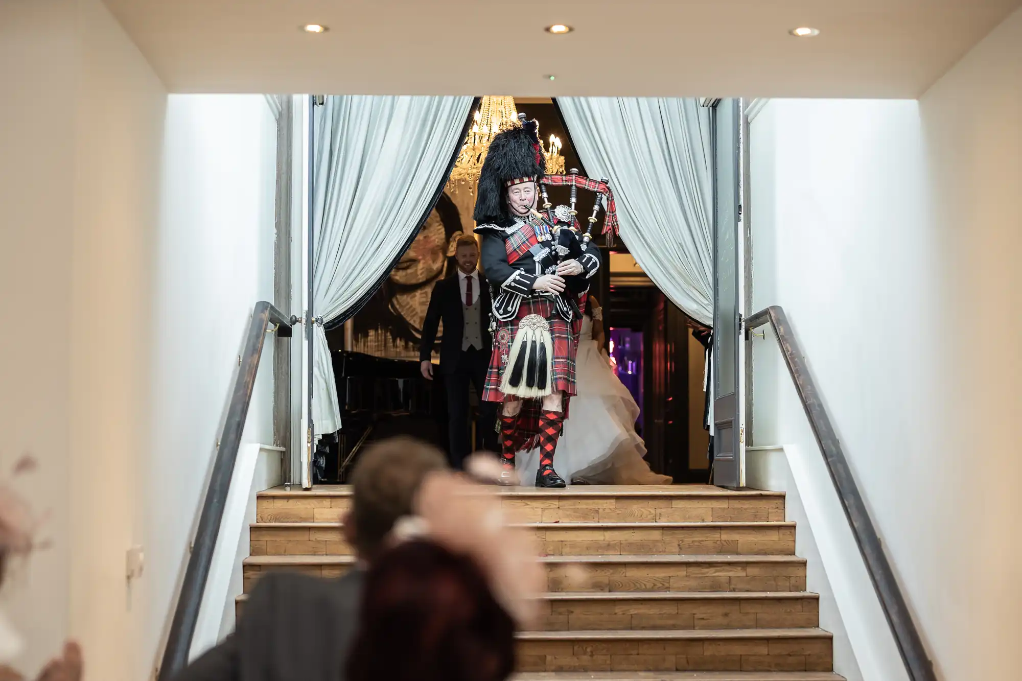 A bagpiper in traditional Scottish attire plays the bagpipes while descending a staircase, followed by two other people in formal wear.