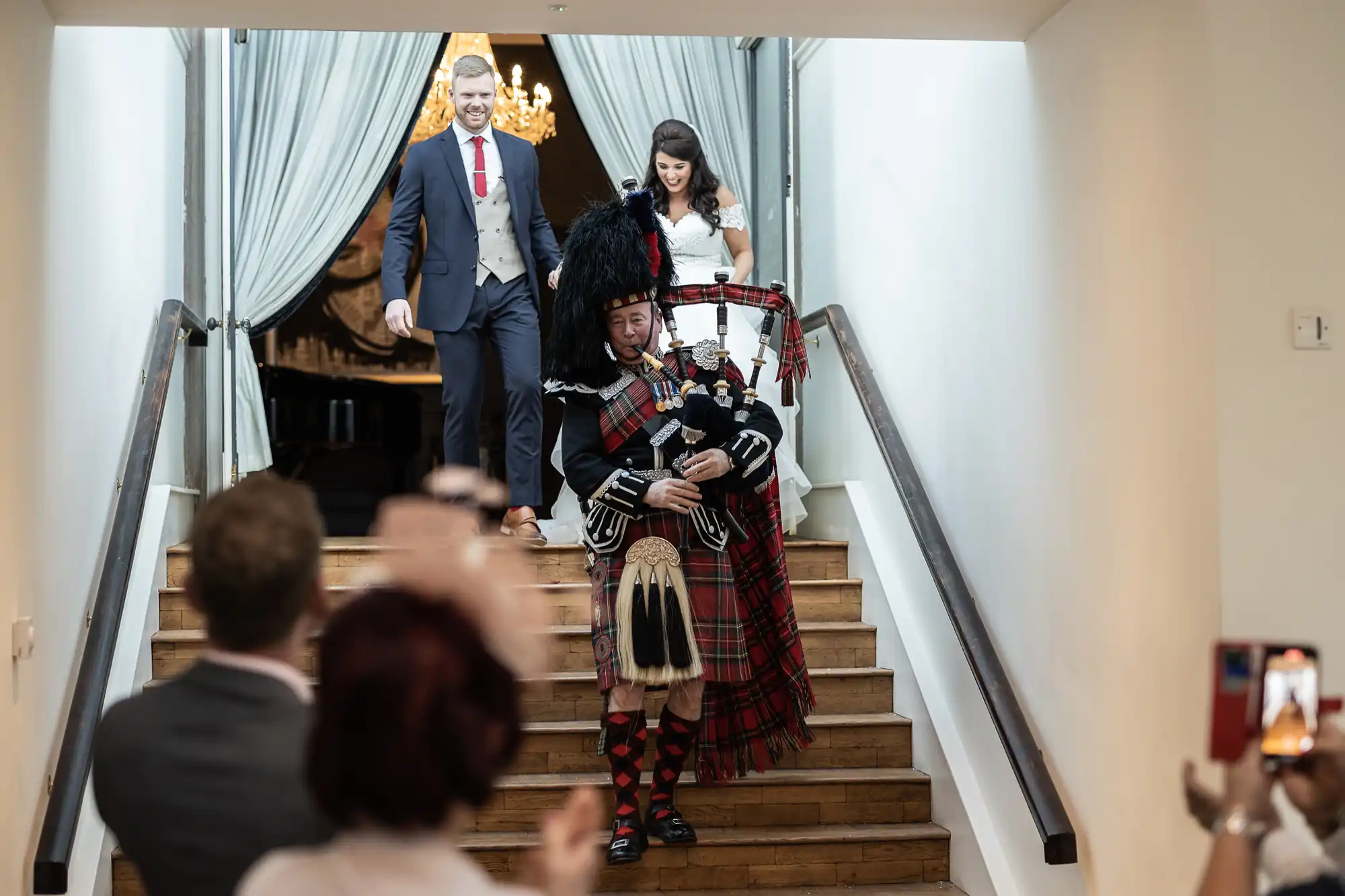 A man and woman descend stairs behind a bagpiper at an event while people admire and take photos.